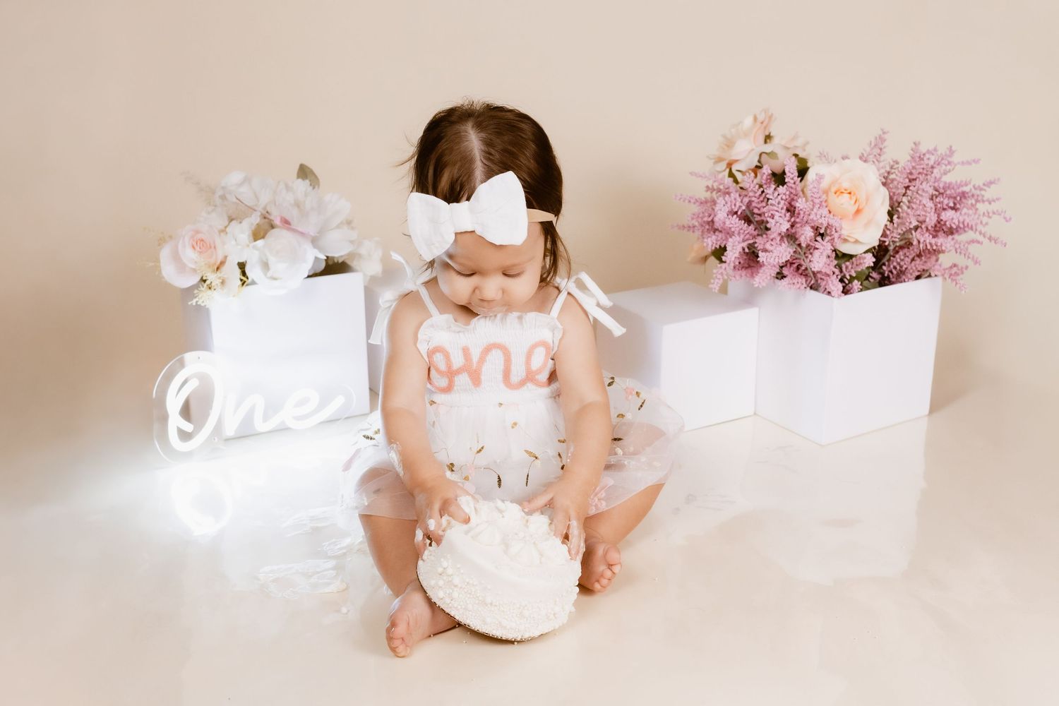 Baby girl in white tutu smashes first birthday cake surrounded by floral arrangements and glowing “one” sign.