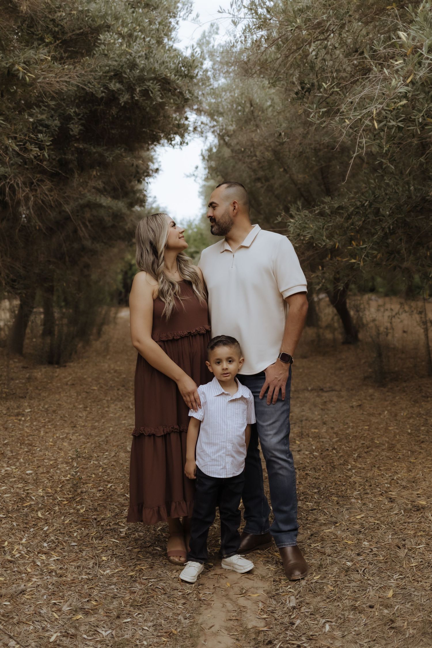 Family of three with young boy standing on a dirt path surrounded by lush green trees.