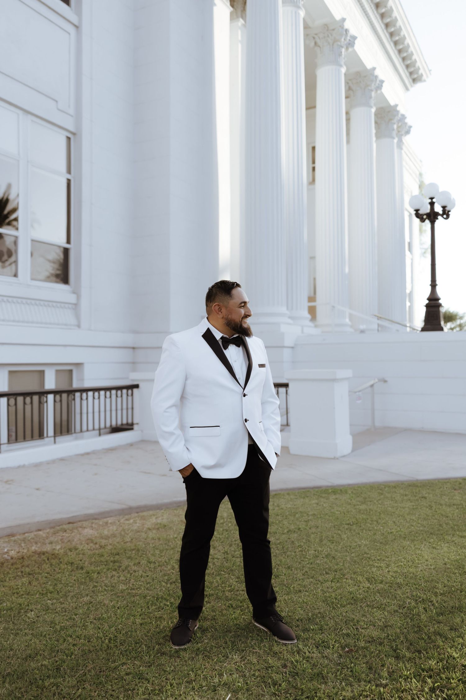 Dapper groom in white tuxedo jacket and black pants standing outside a columned building.