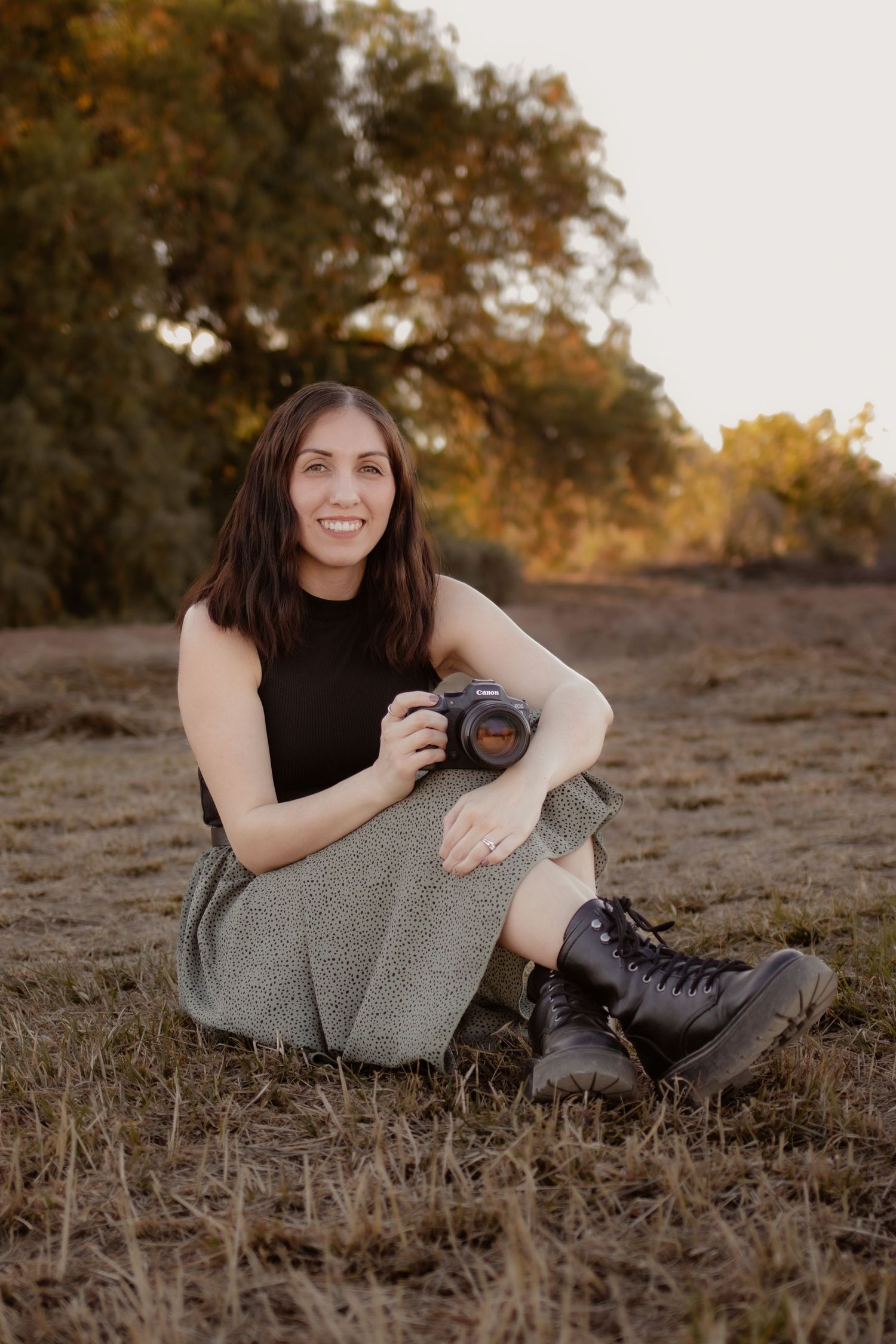 Smiling female photographer sitting in a field holding a DSLR camera at golden hour.