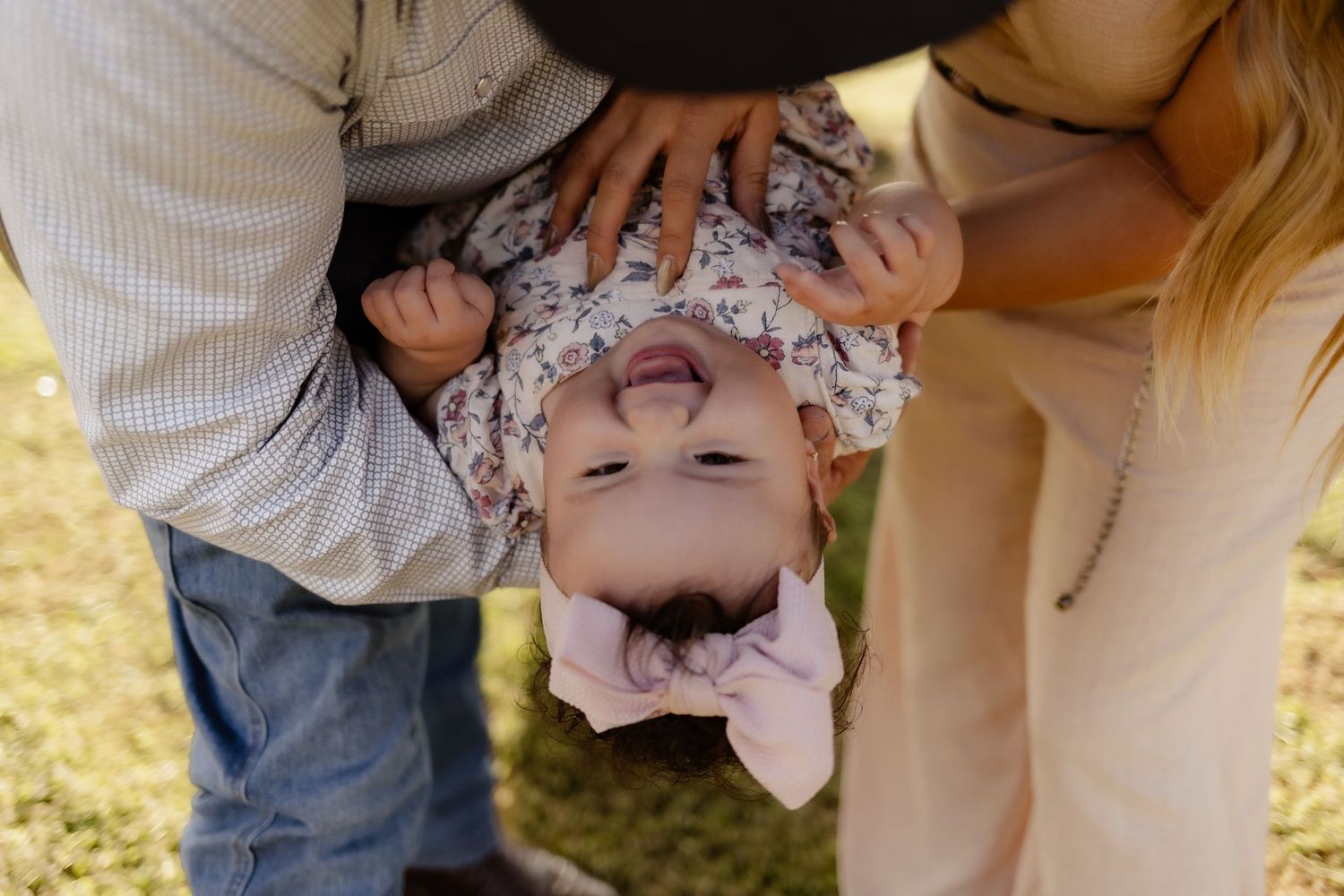 Two parents playfully hold their laughing baby upside down on a sunny green lawn.