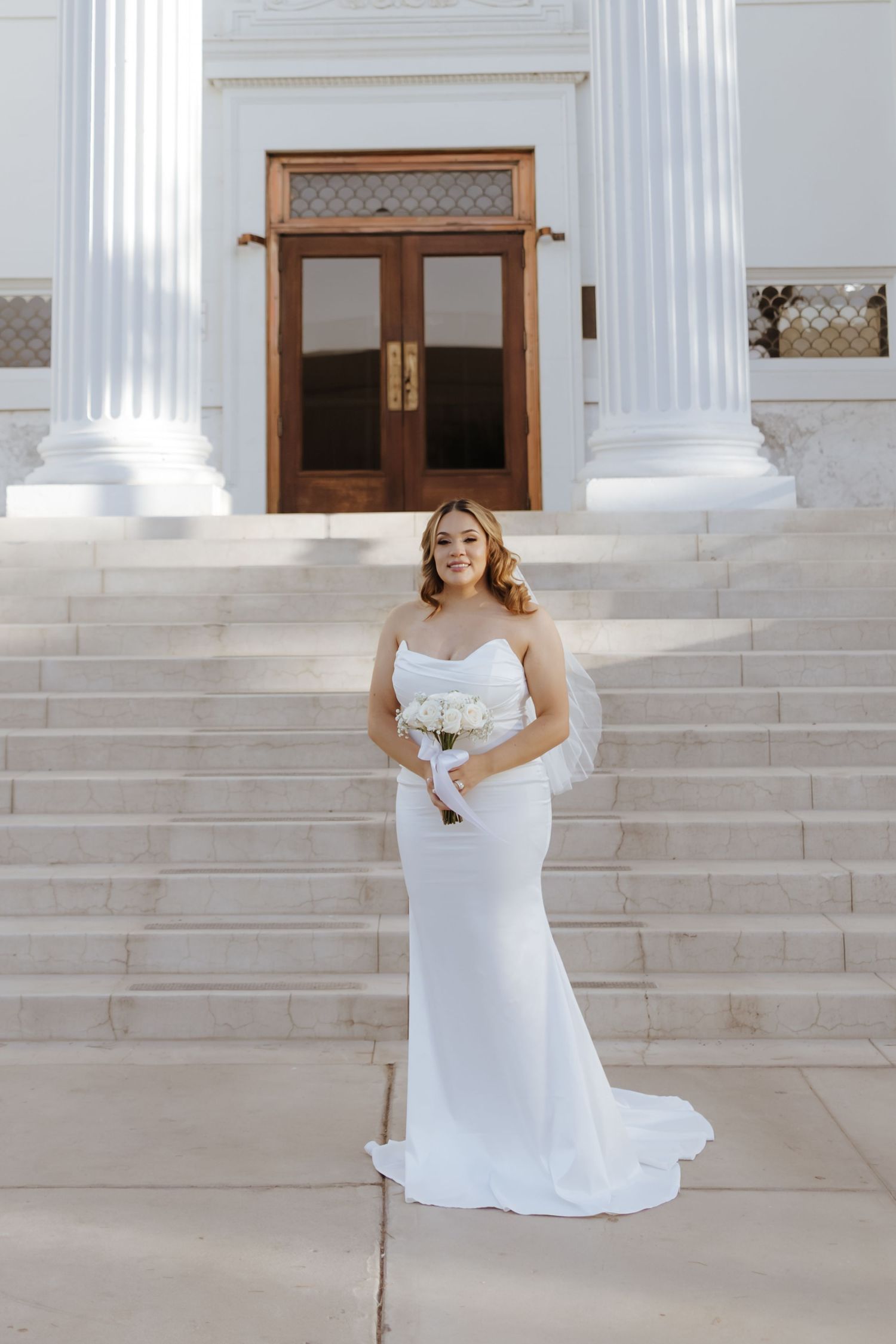 Bride in sleek strapless white gown holding bouquet on marble steps of grand building.