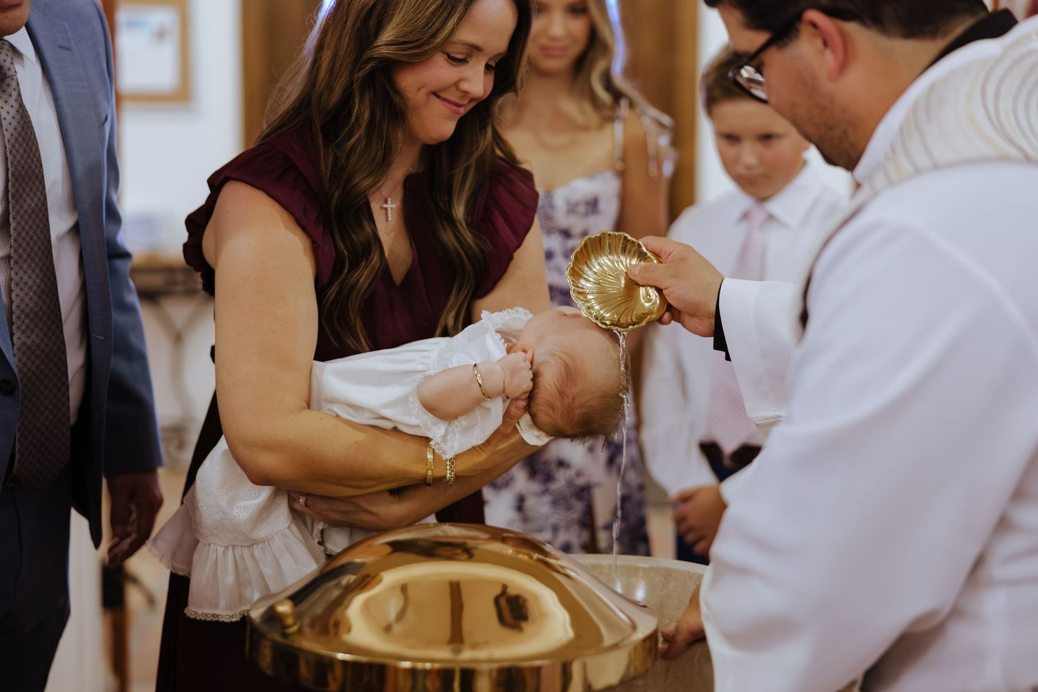 Woman in burgundy dress holding infant during Catholic baptism ceremony at church font.