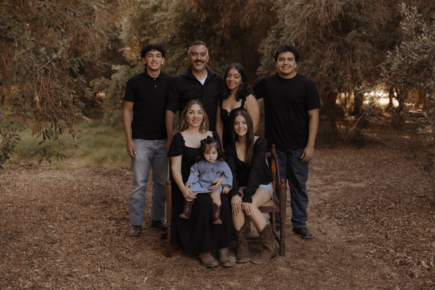 Family of seven posing together outdoors among trees in coordinating dark outfits for portrait.