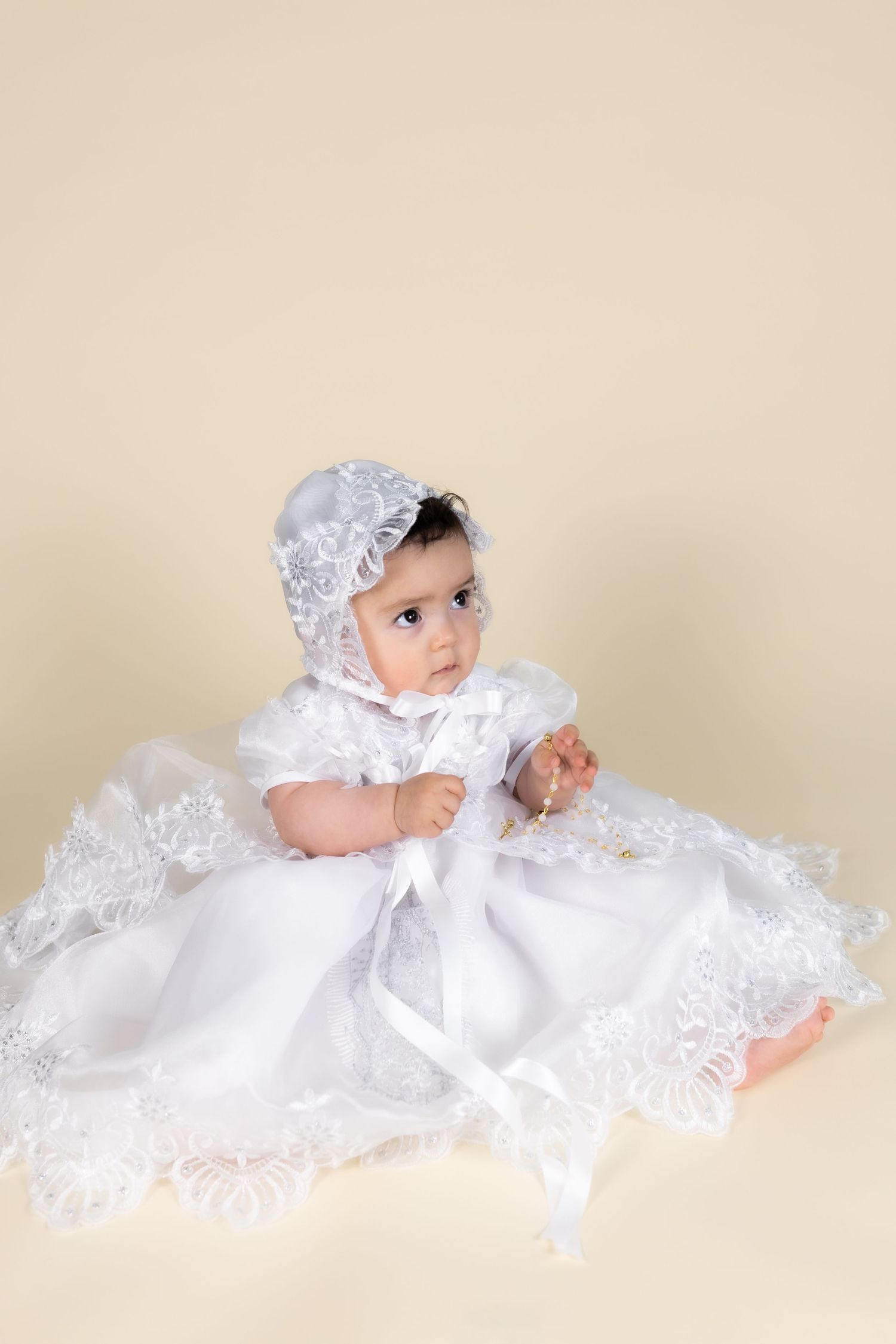 Baby girl in white lace christening gown and bonnet sitting on cream background.