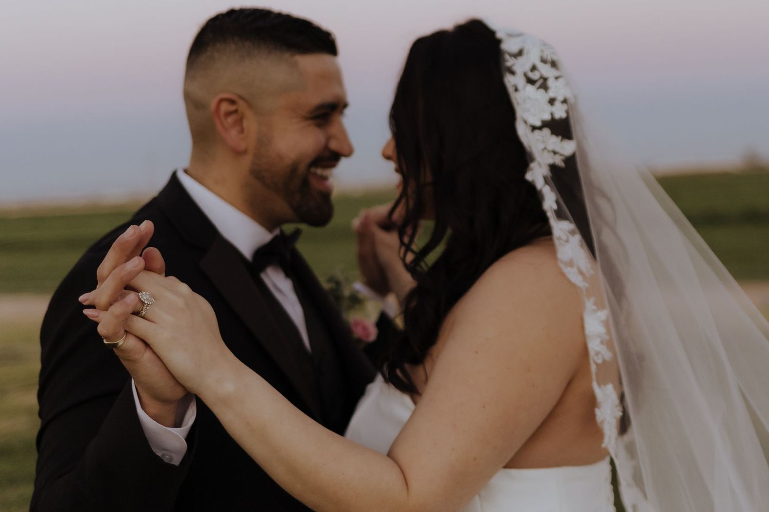 Bride and groom share a romantic dance outdoors at sunset, groom in black suit with floral boutonniere.