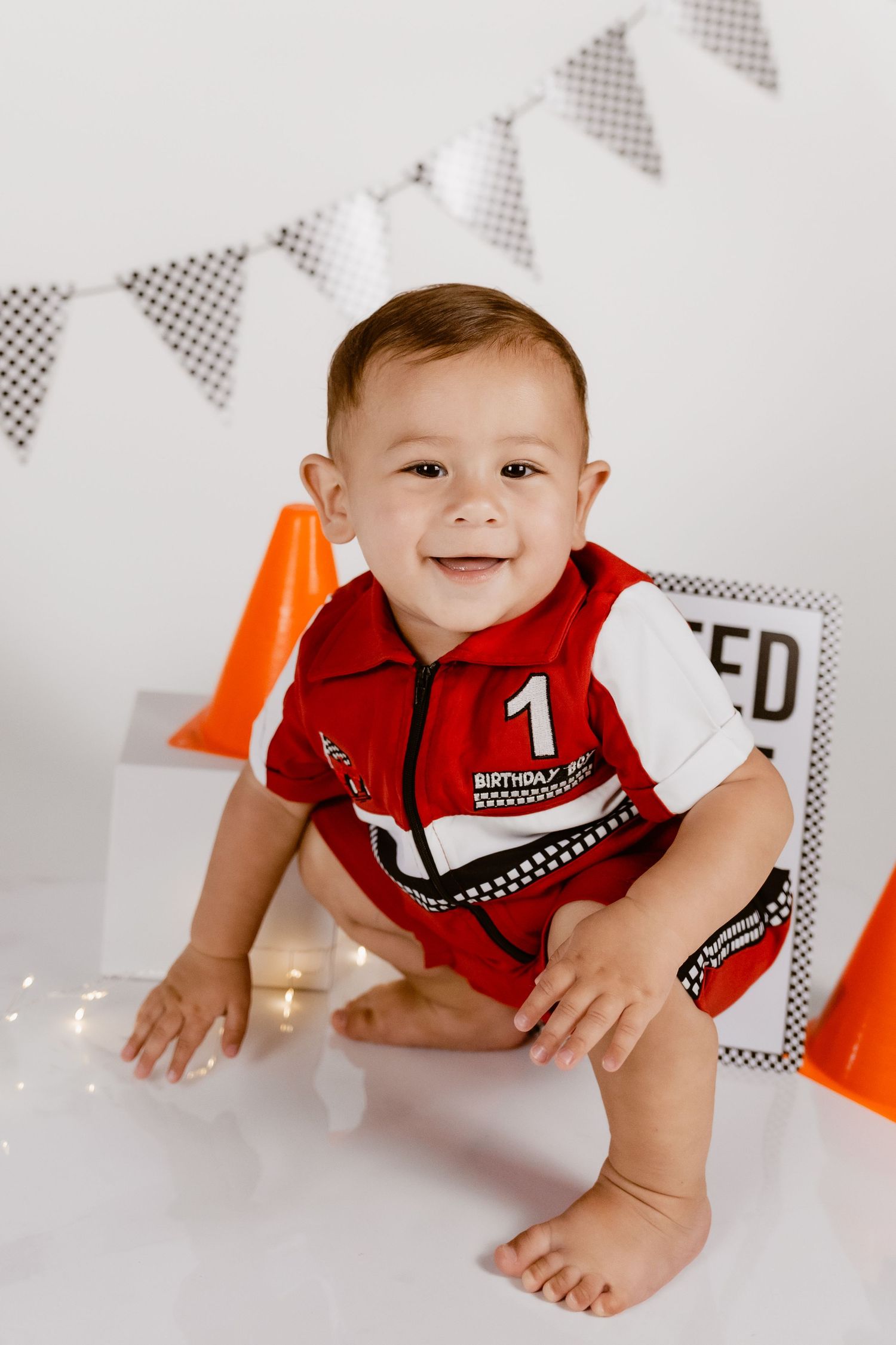 Smiling baby boy in red racing driver outfit sitting on white floor with orange cones and bunting for first birthday.
