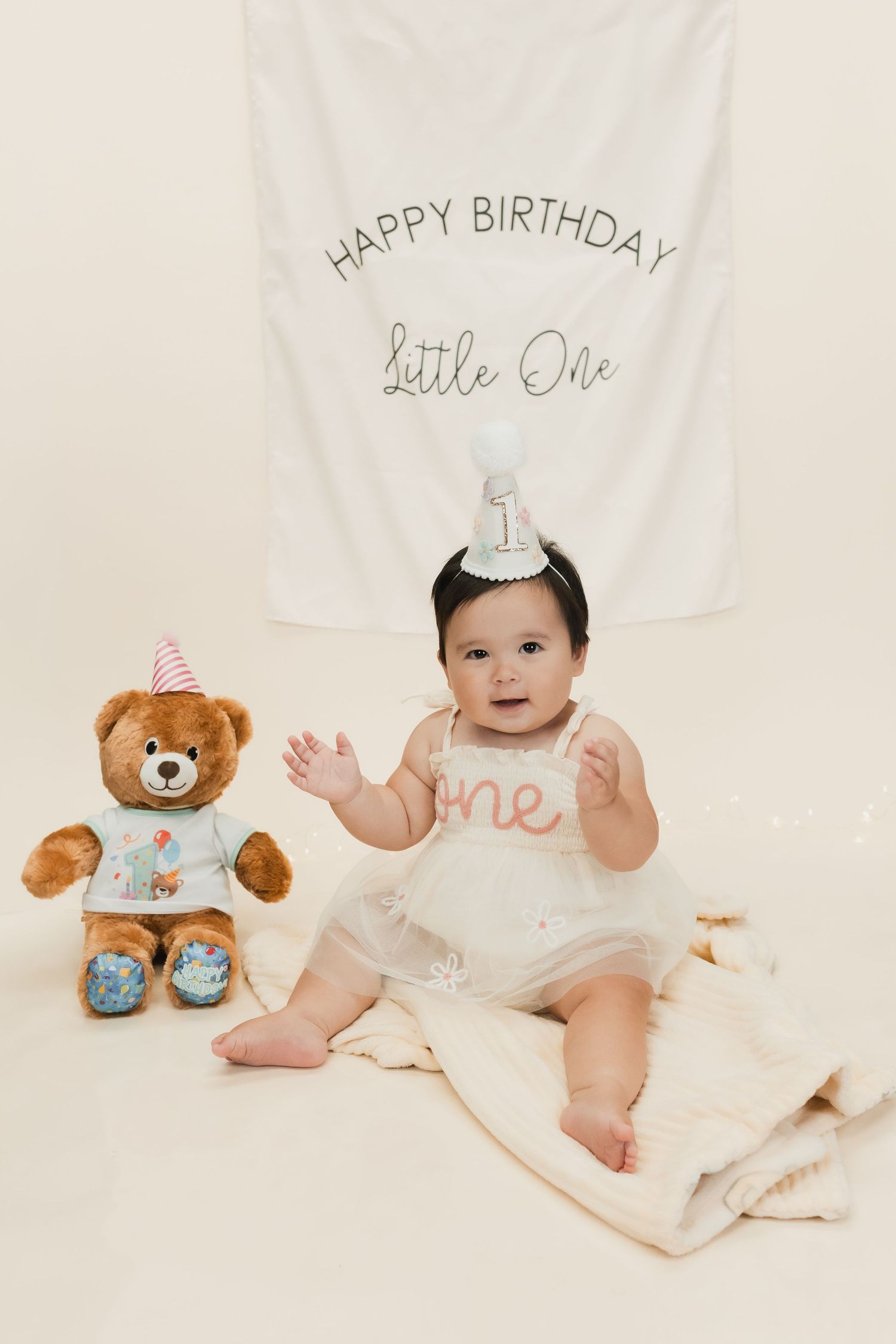 Baby girl in white tutu sits beside teddy bear under Happy Birthday Little One banner for first birthday celebration.
