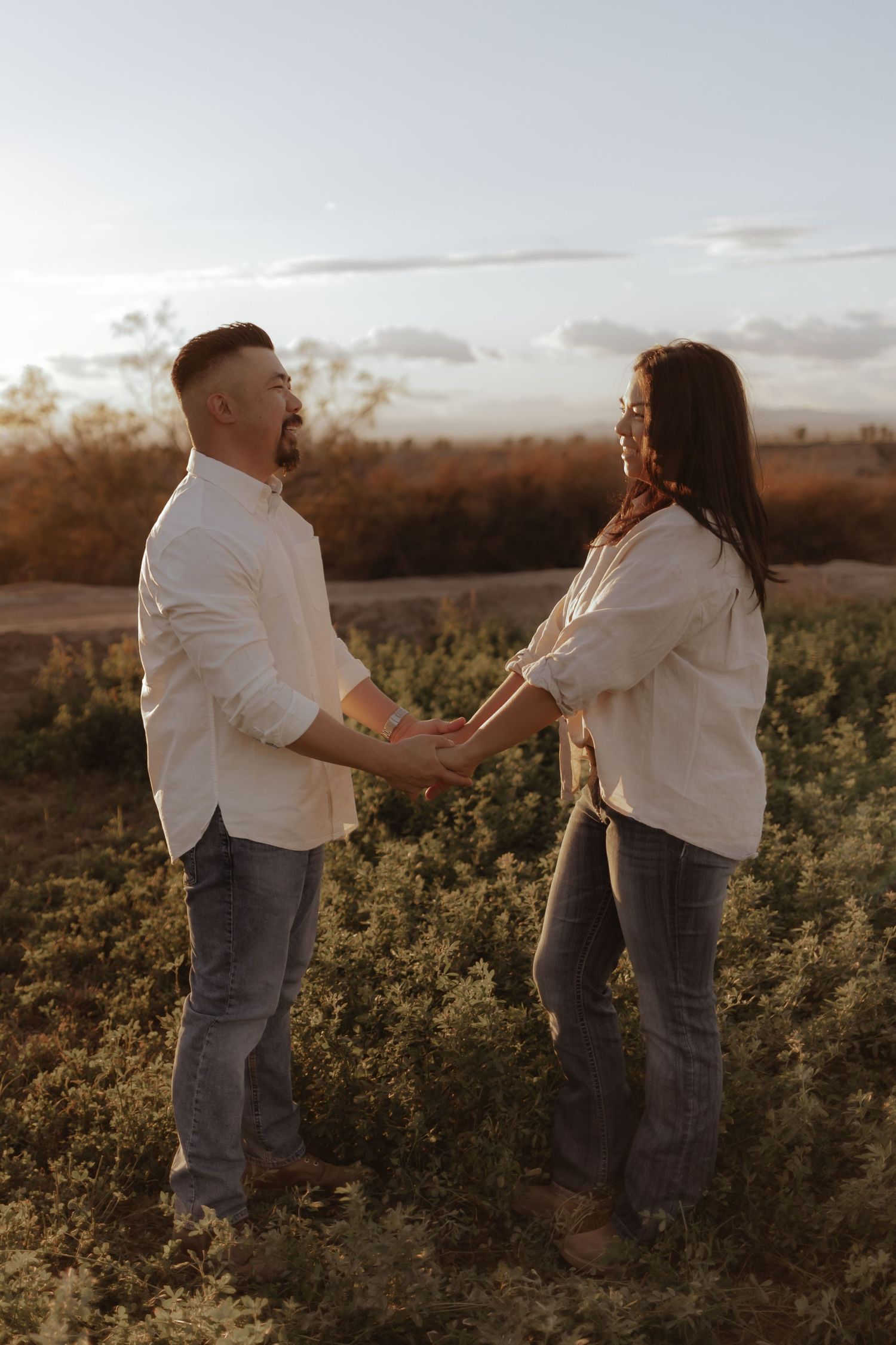 Couple in white shirts holds hands facing each other in golden sunset field.