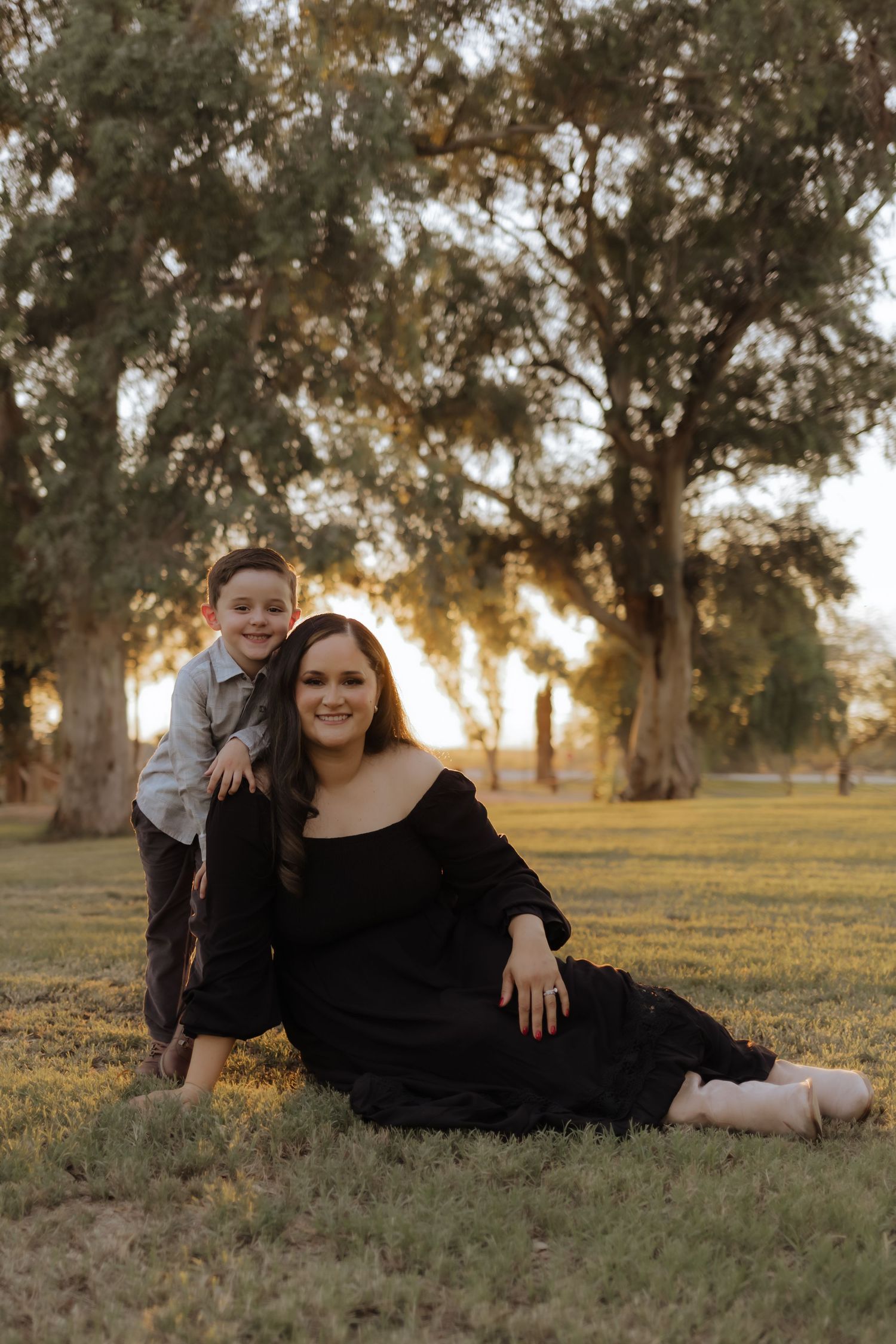 Mother in black dress sits on grass with young son leaning on her at sunset.