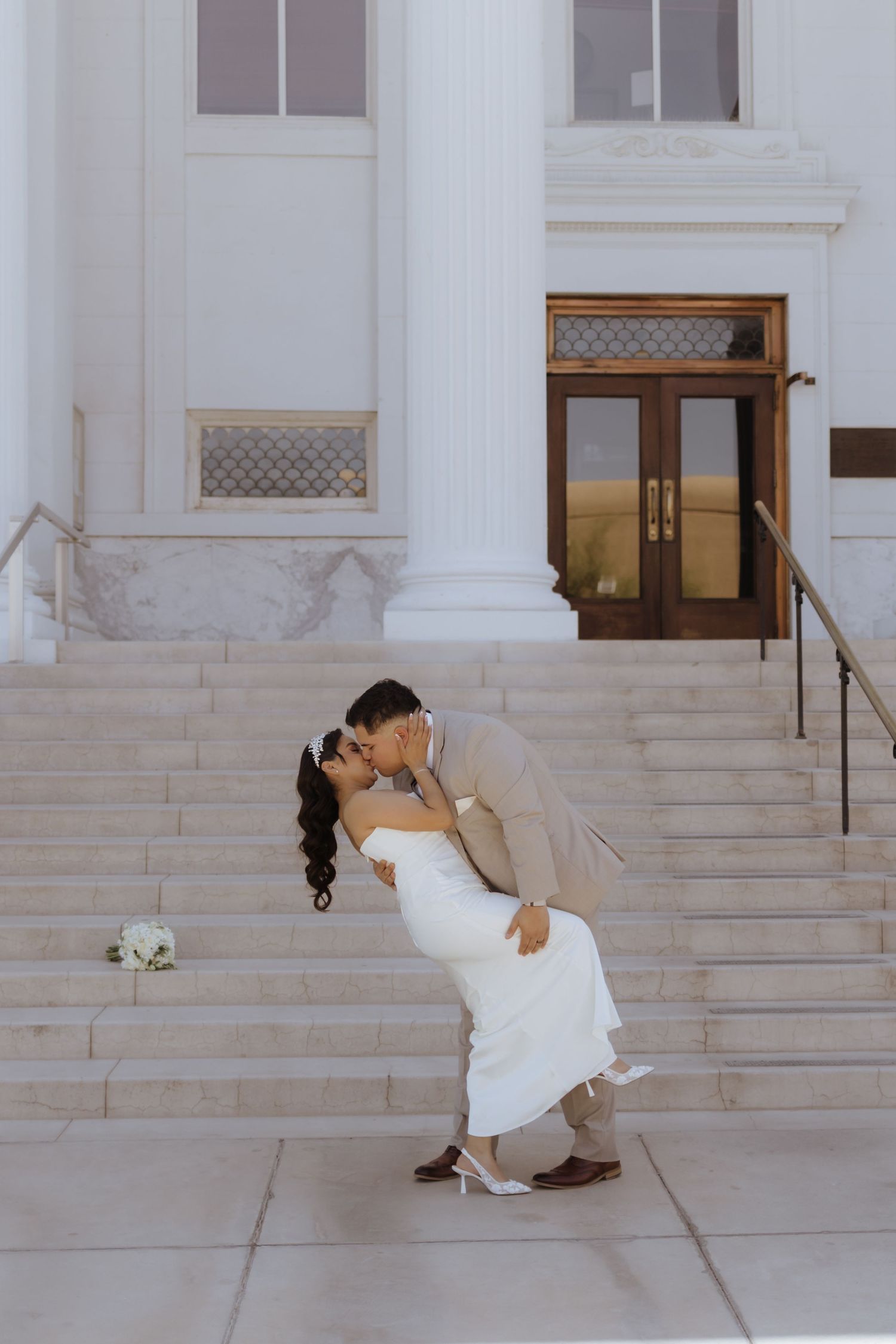 Groom dips bride for a kiss on the steps of a grand white classical building in bright sunlight.