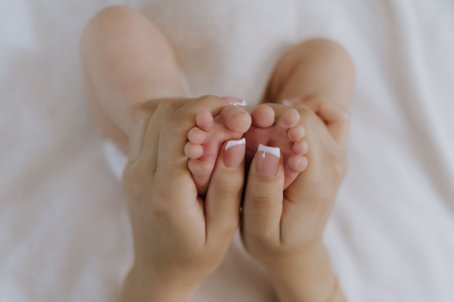 Adult hands gently cradling tiny newborn baby feet on a soft white background.