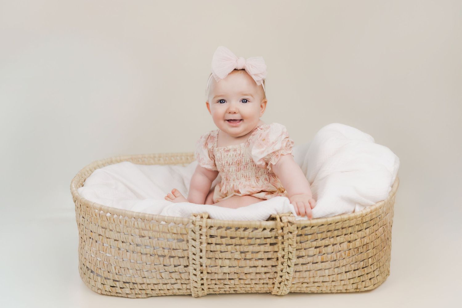 Smiling baby girl in pink dress and bow headband sitting in a woven Moses basket with white blanket.
