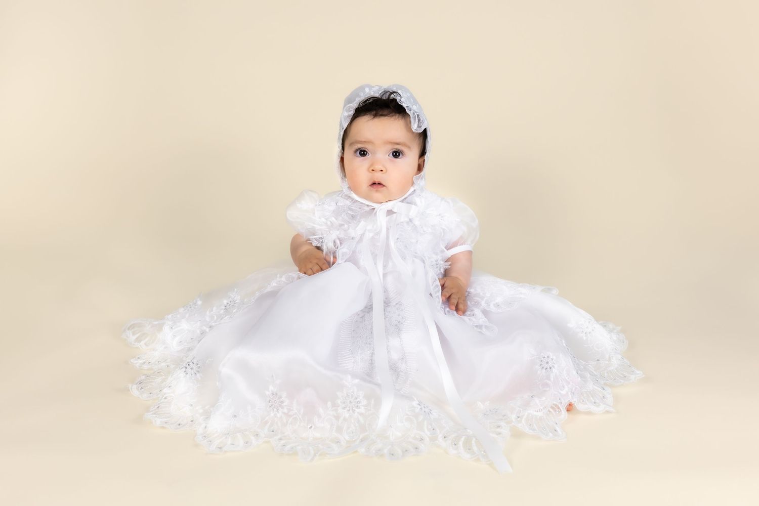 Baby girl in white lace christening gown and bonnet sitting gracefully on a cream background.