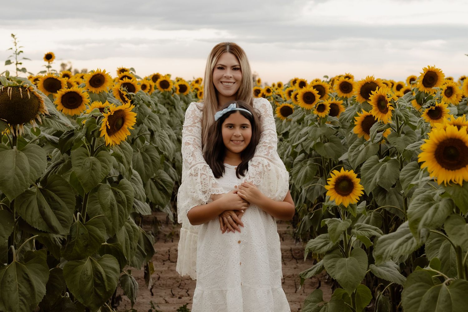 Two girls in white dresses embracing in a vibrant sunflower field at dusk.