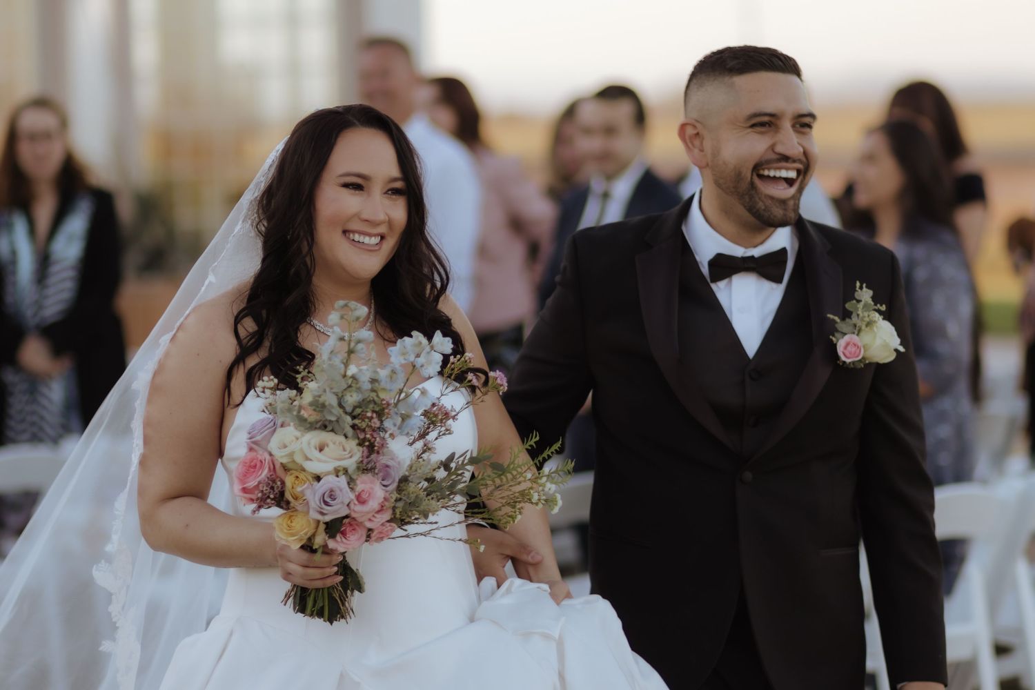 Smiling bride and groom walk down the aisle after their outdoor wedding ceremony, bride holding floral bouquet.