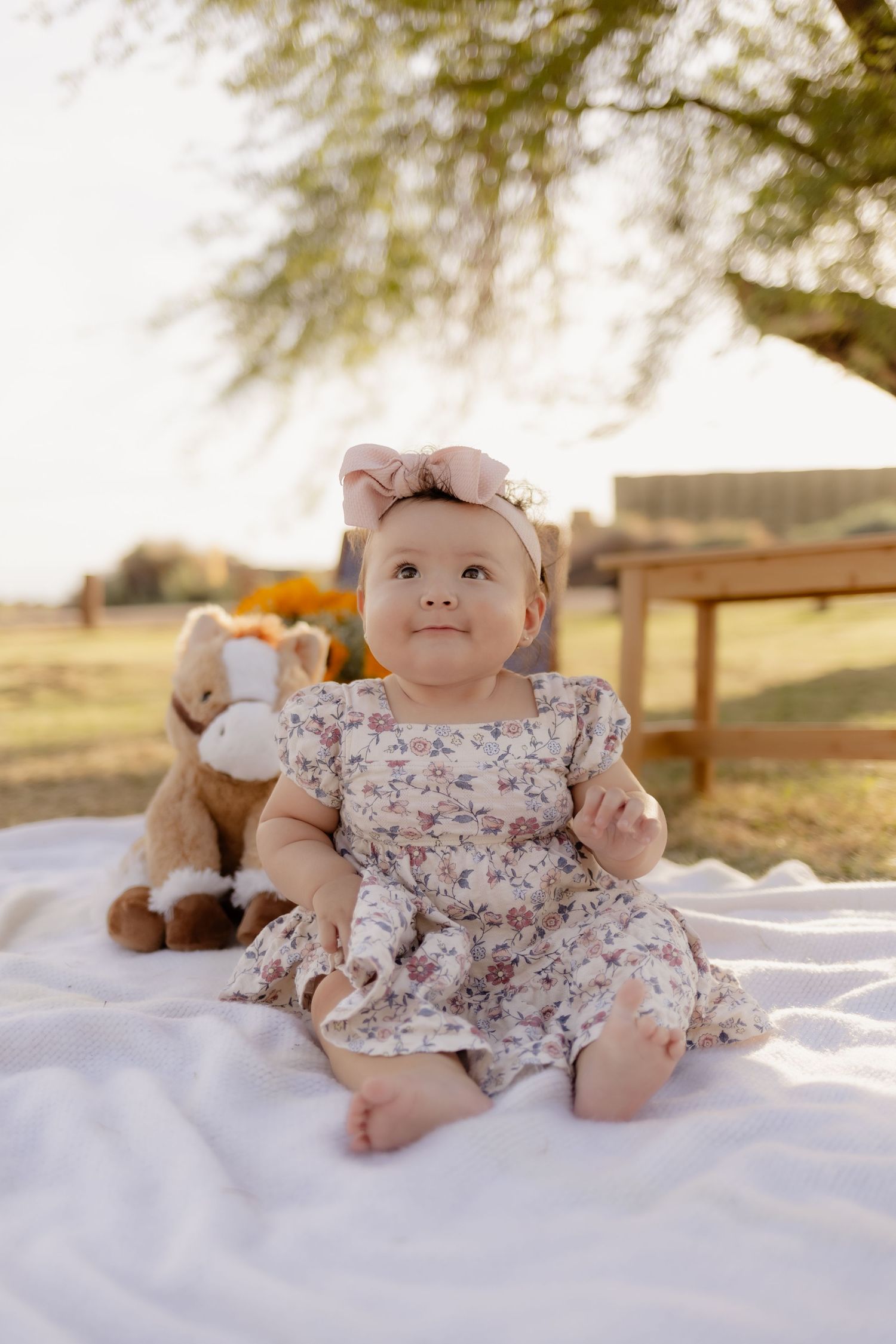 An adorable baby in a floral dress and pink bow headband sits on a white blanket with stuffed animals.