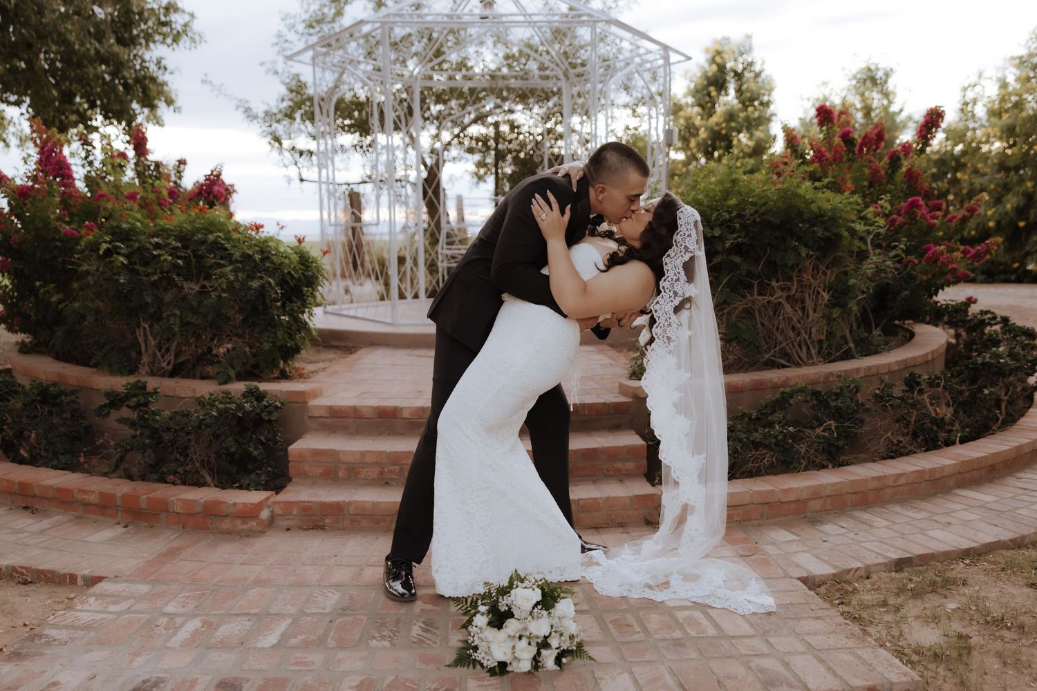 Bride and groom share a romantic dip kiss in a garden wedding venue surrounded by red roses and brick steps.