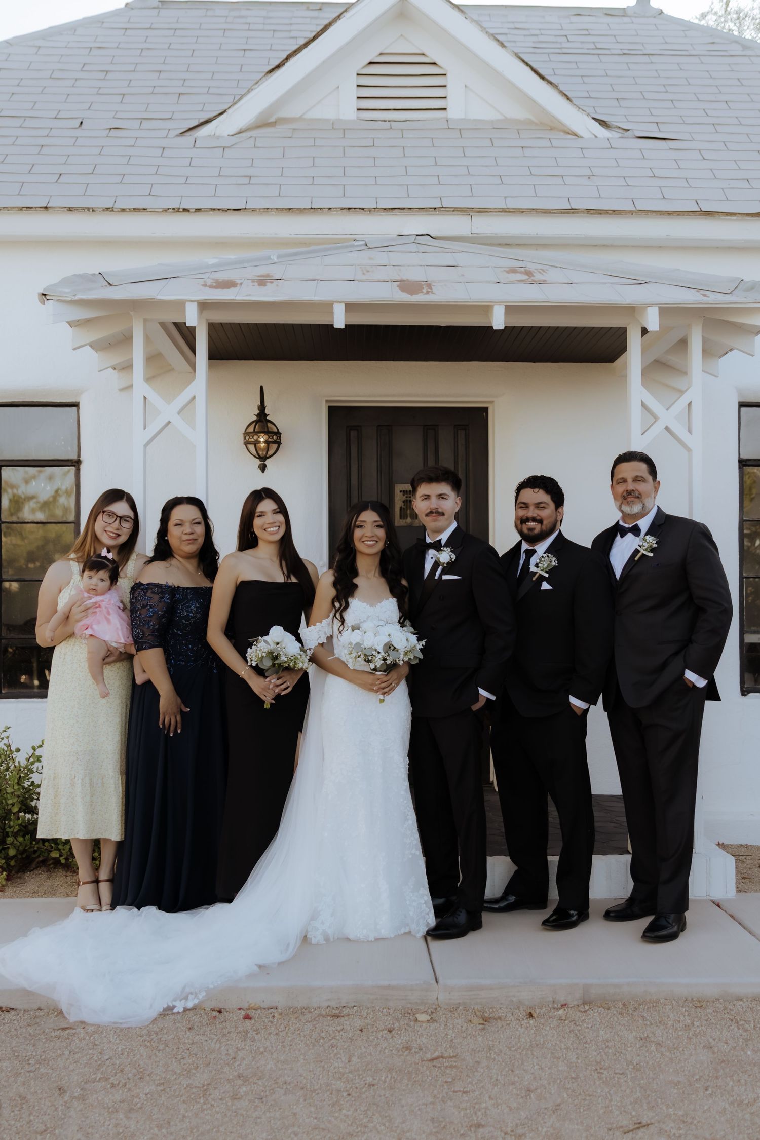 Wedding party poses outside white chapel venue with bride in lace gown and groomsmen in black tuxedos.