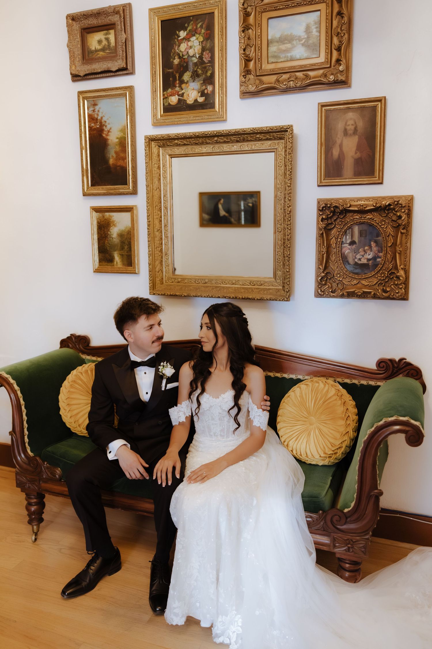 Bride and groom sitting together on a green velvet antique sofa surrounded by ornate gold-framed paintings.