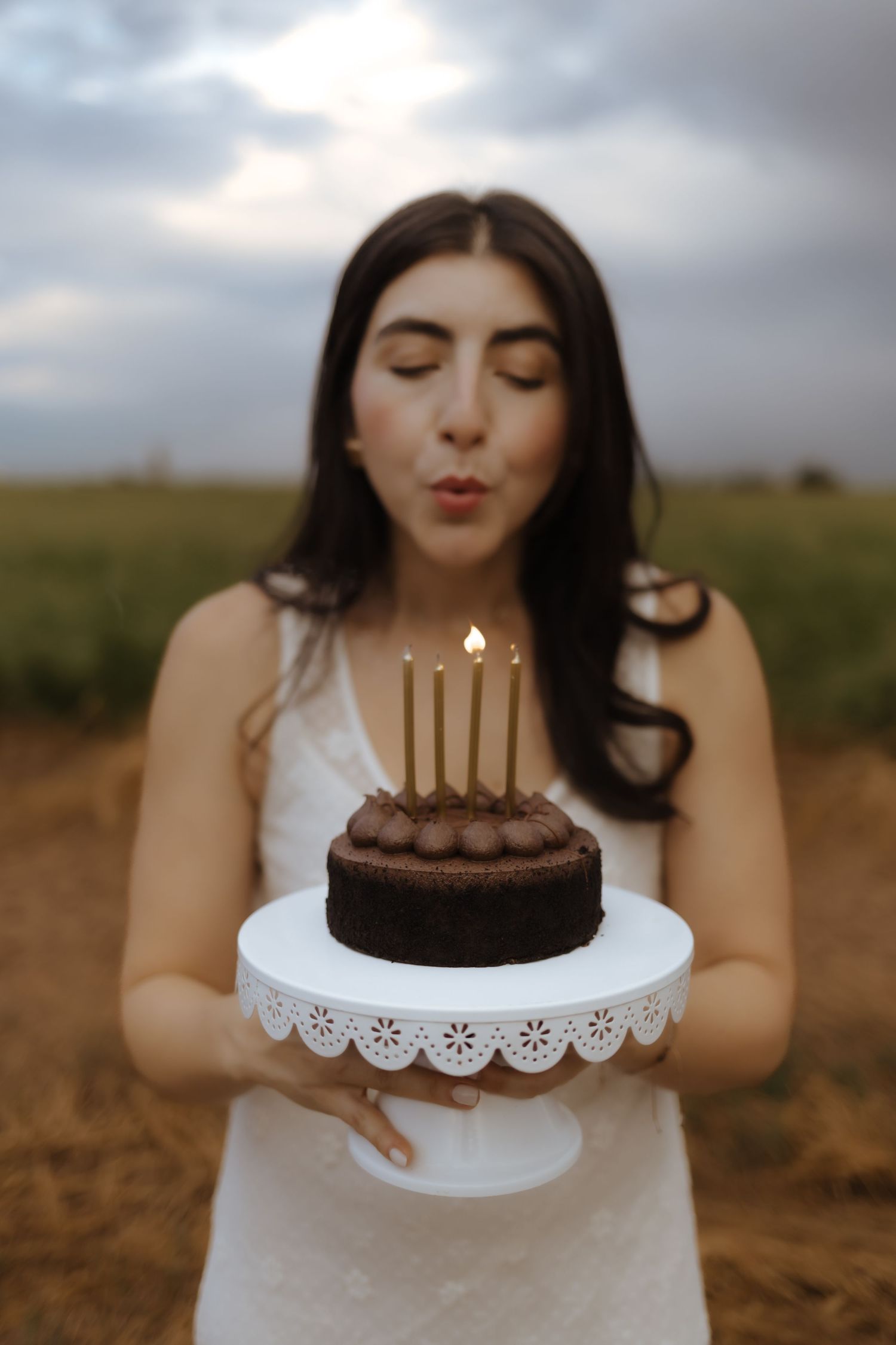 Woman in white dress blowing out gold candles on a chocolate birthday cake outdoors.