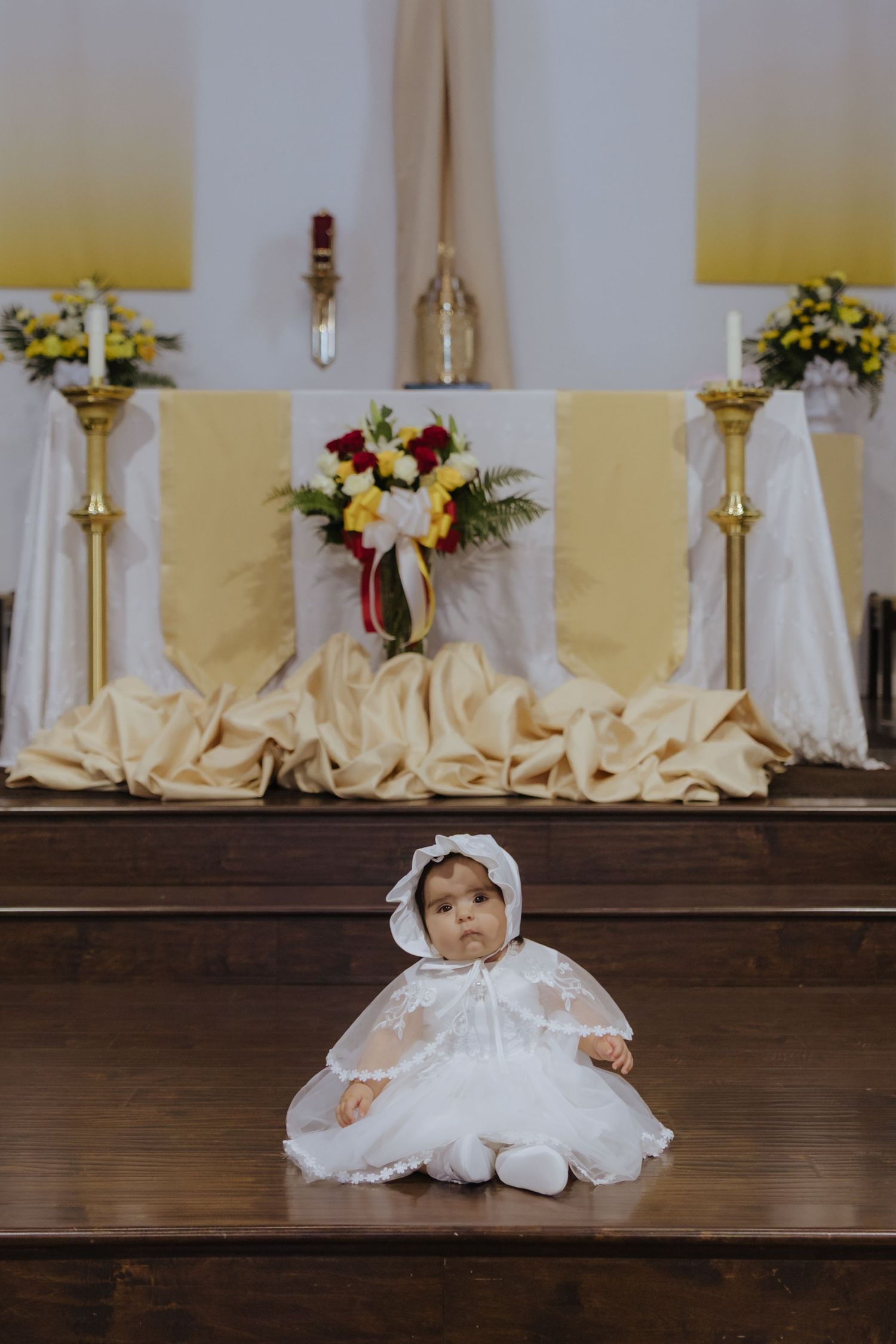 Baby in white baptism gown and bonnet sitting on church altar steps during ceremony.