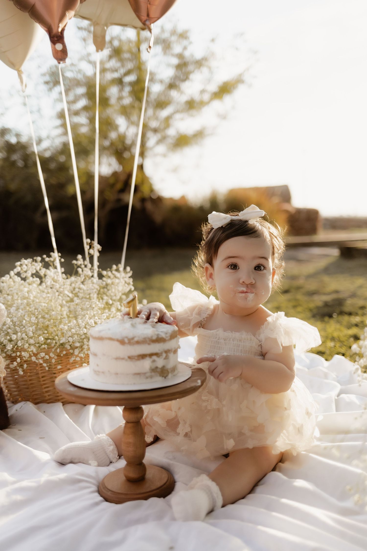 Baby girl in white dress touching first birthday cake outdoors with balloons above.