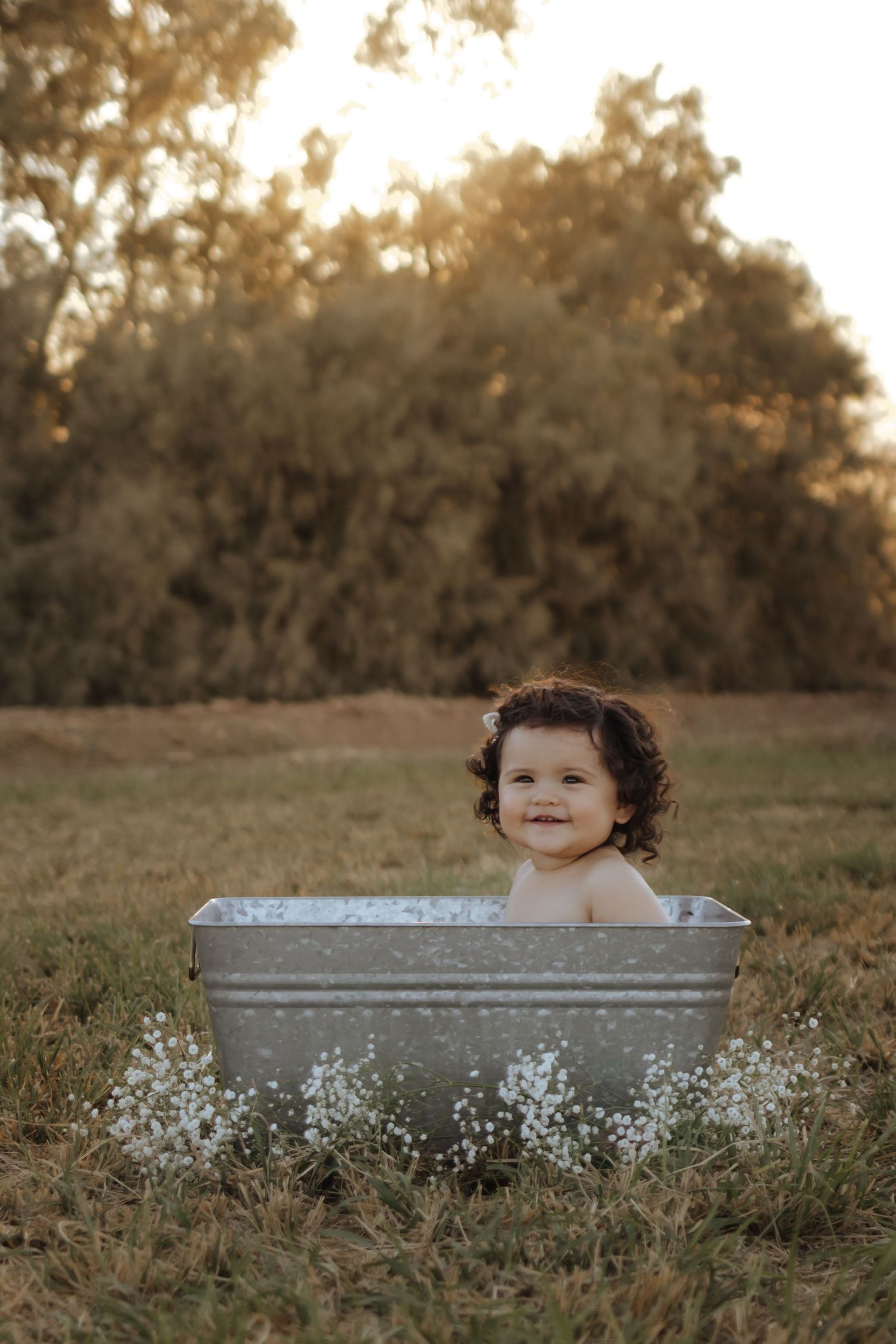 Curly-haired toddler smiles sitting in galvanized tub outdoors at golden hour.