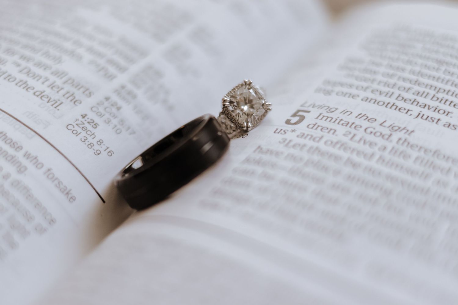 Diamond engagement ring and black wedding band rest open on a Bible page in an elegant wedding detail photo.