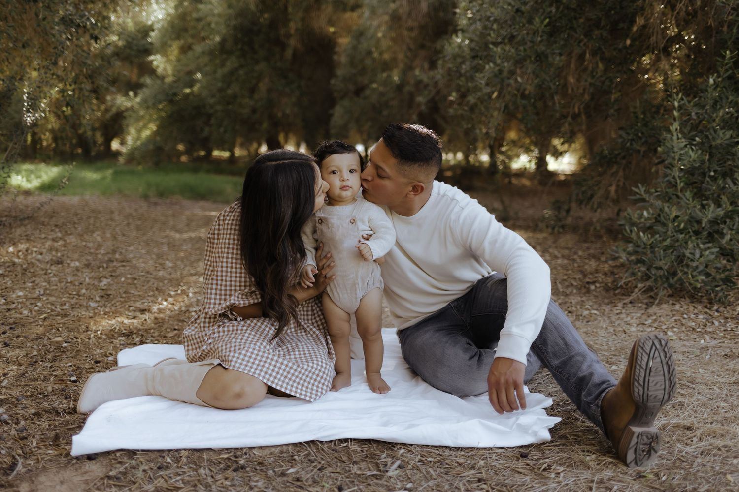 A young family sits on a white blanket outdoors, parents kissing their baby on the cheeks in a serene forest setting.