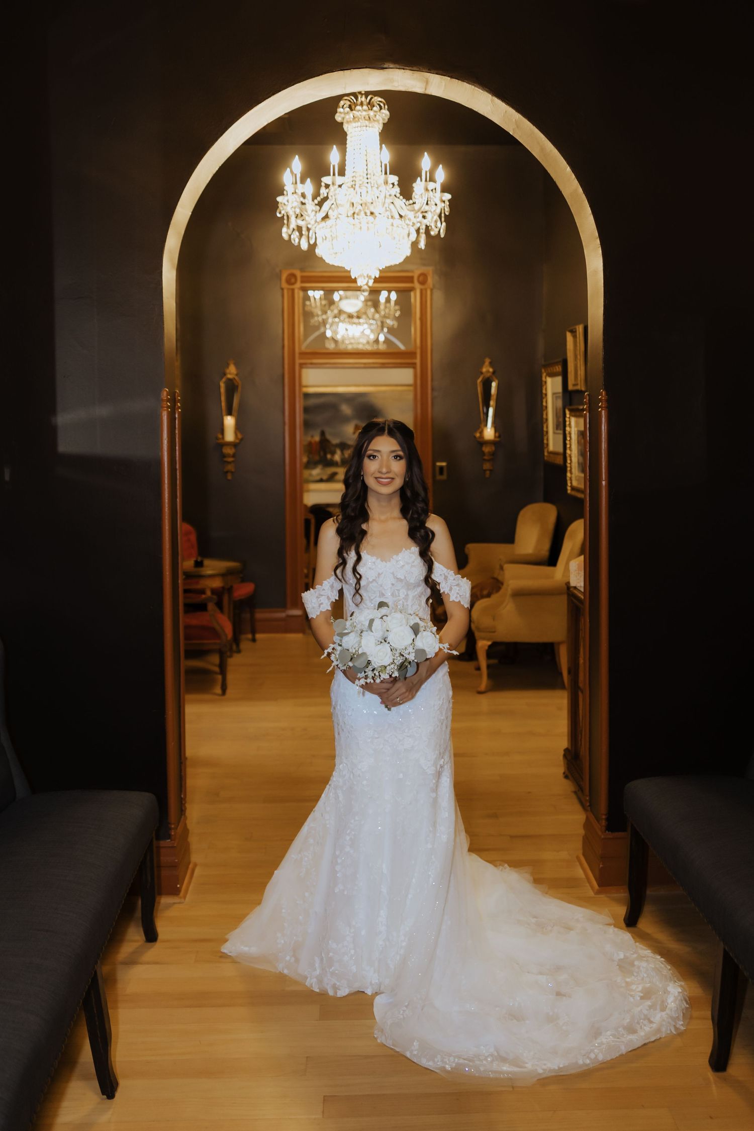 Bride in off-shoulder lace gown holding white bouquet stands under gold arch with chandelier.