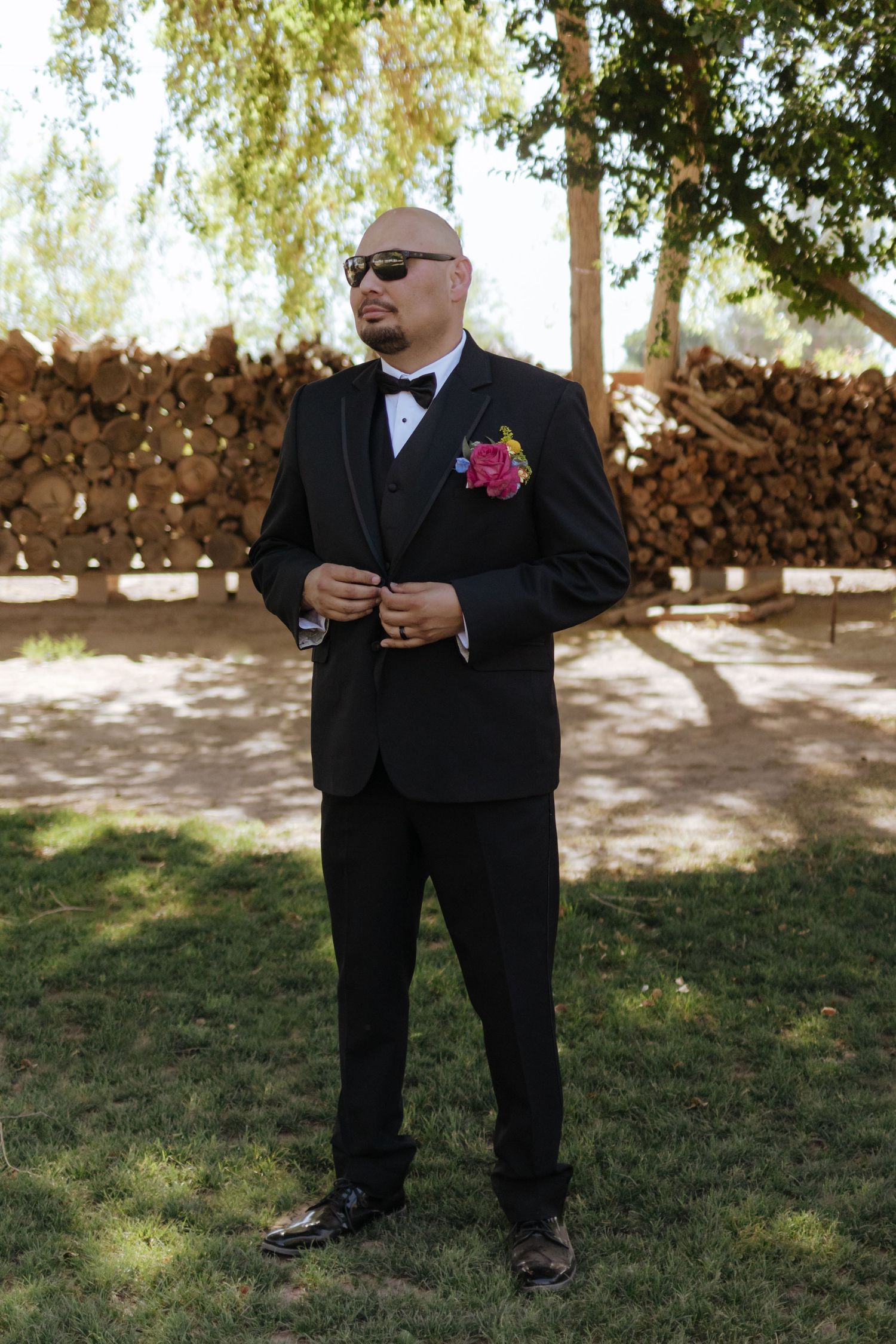 Bald groom in black tuxedo with colorful boutonniere and sunglasses standing outdoors near stacked logs.