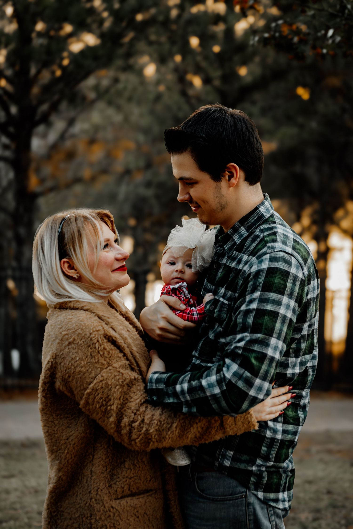 A young family with a blonde woman in a tan coat and a man in plaid holding their baby outdoors in autumn.