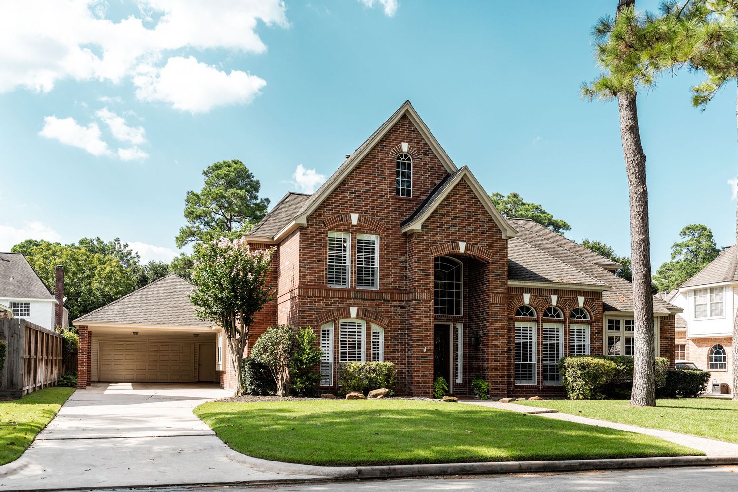 Brick two-story home with attached garage, manicured lawn, and tall pine trees under a bright blue sky.