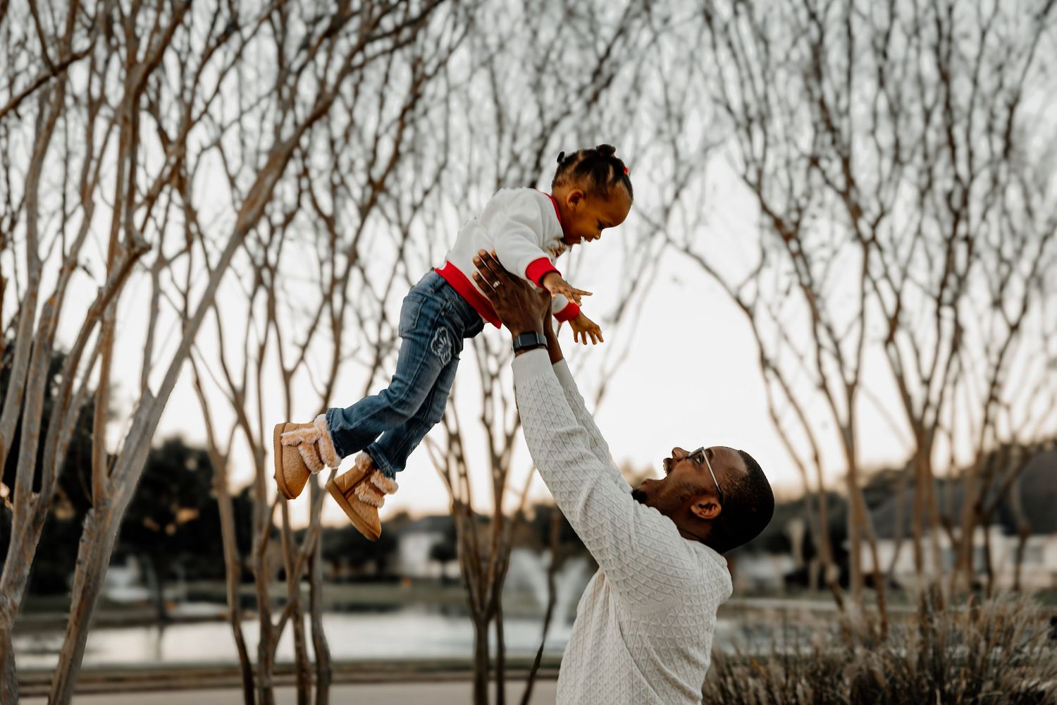 Father joyfully lifting his young child into the air near bare winter trees.