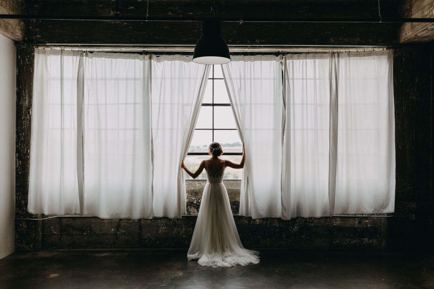Bride in wedding gown standing before large curtained windows in rustic venue.