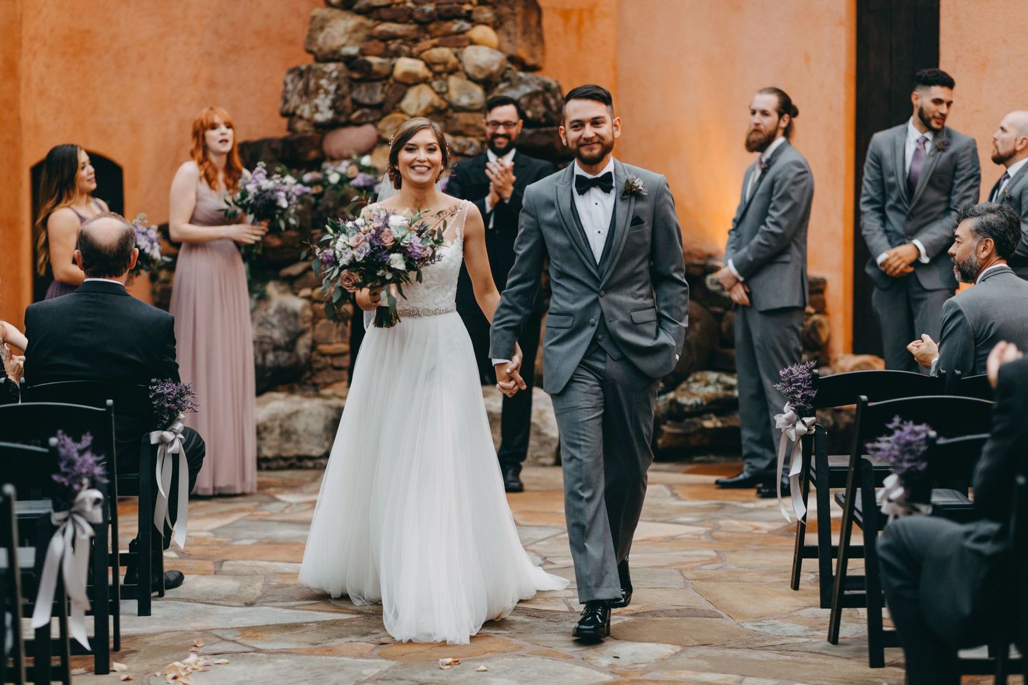 Smiling newlyweds walking down aisle in rustic stone venue with purple floral bouquet.