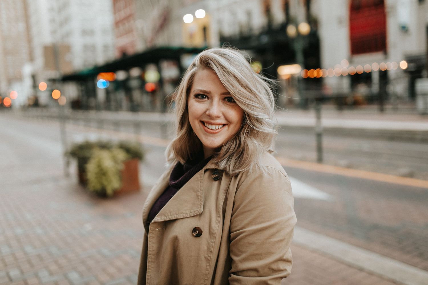 Smiling blonde woman in tan trench coat on city street with bokeh lights background.