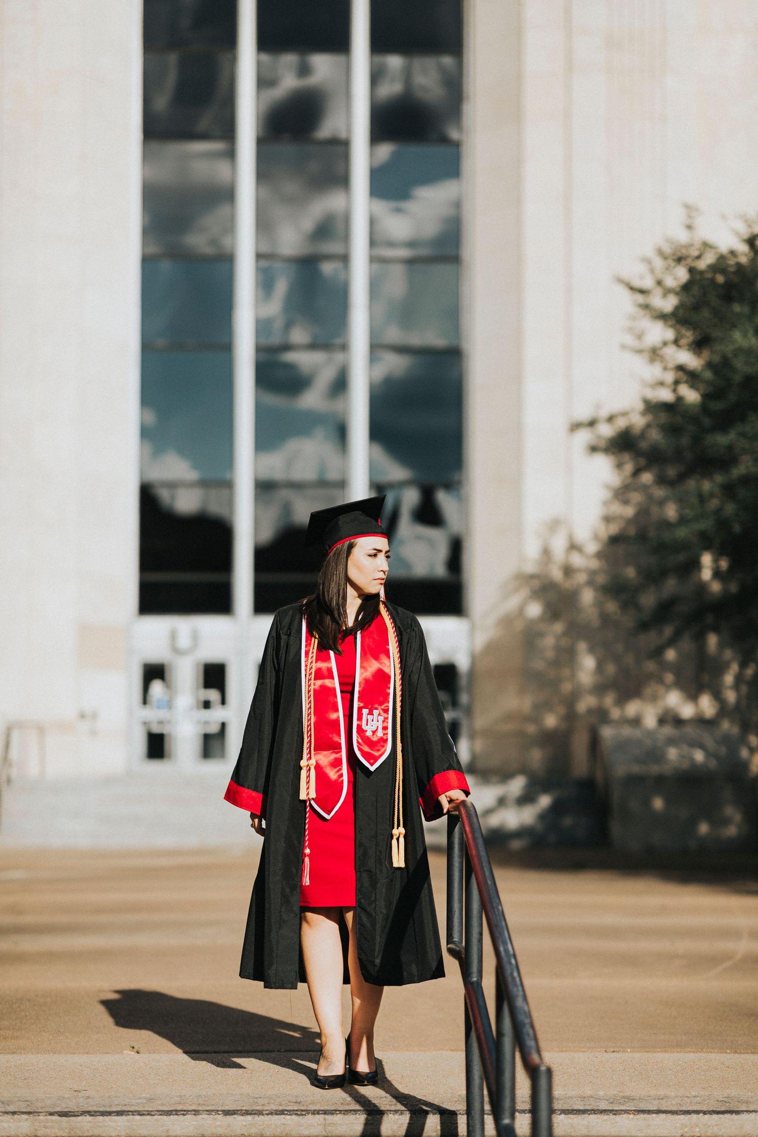 Female graduate in black cap and gown with red stole poses on steps outside university building.