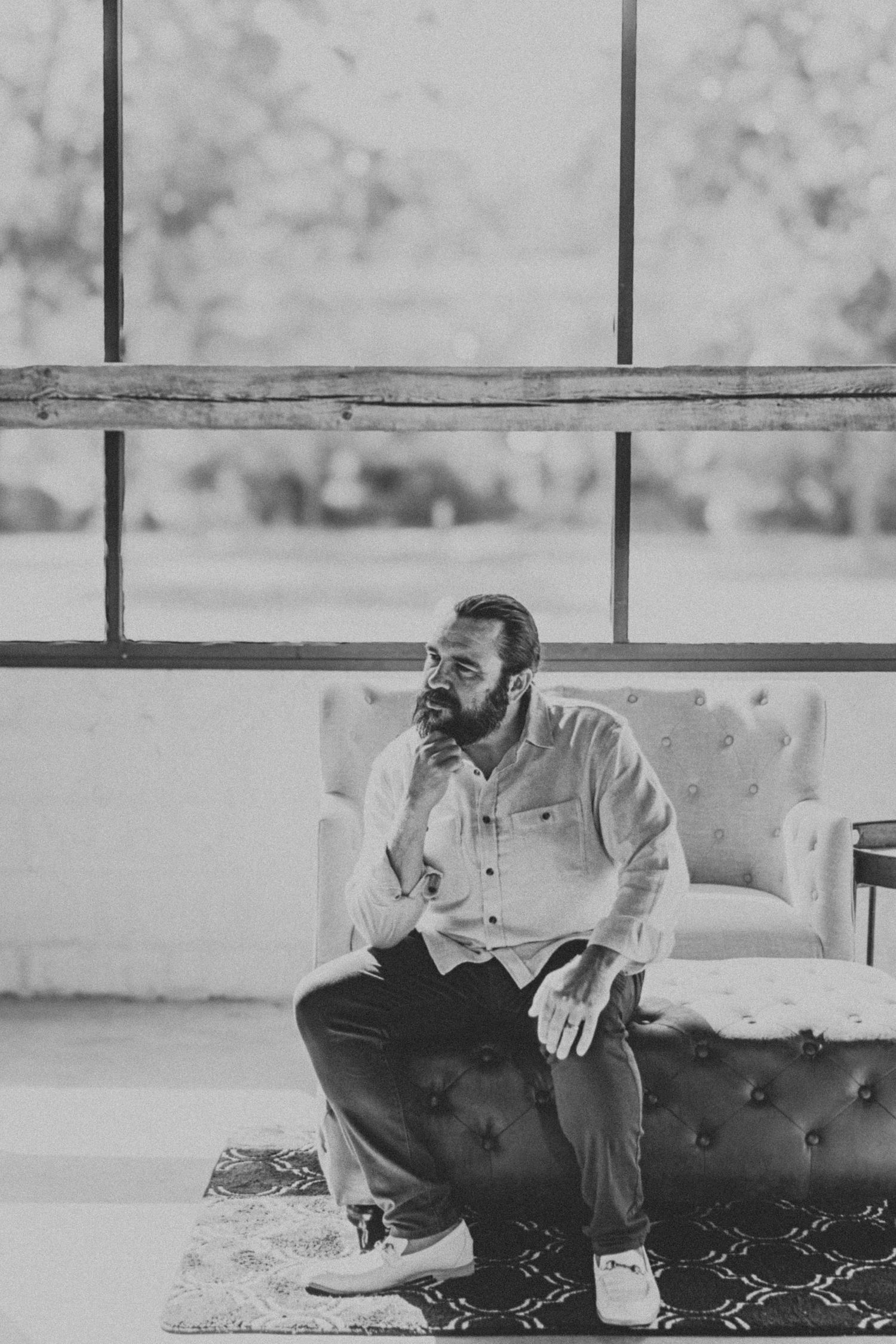 Bearded man in casual shirt sits thoughtfully on tufted sofa near large windows in black and white portrait.