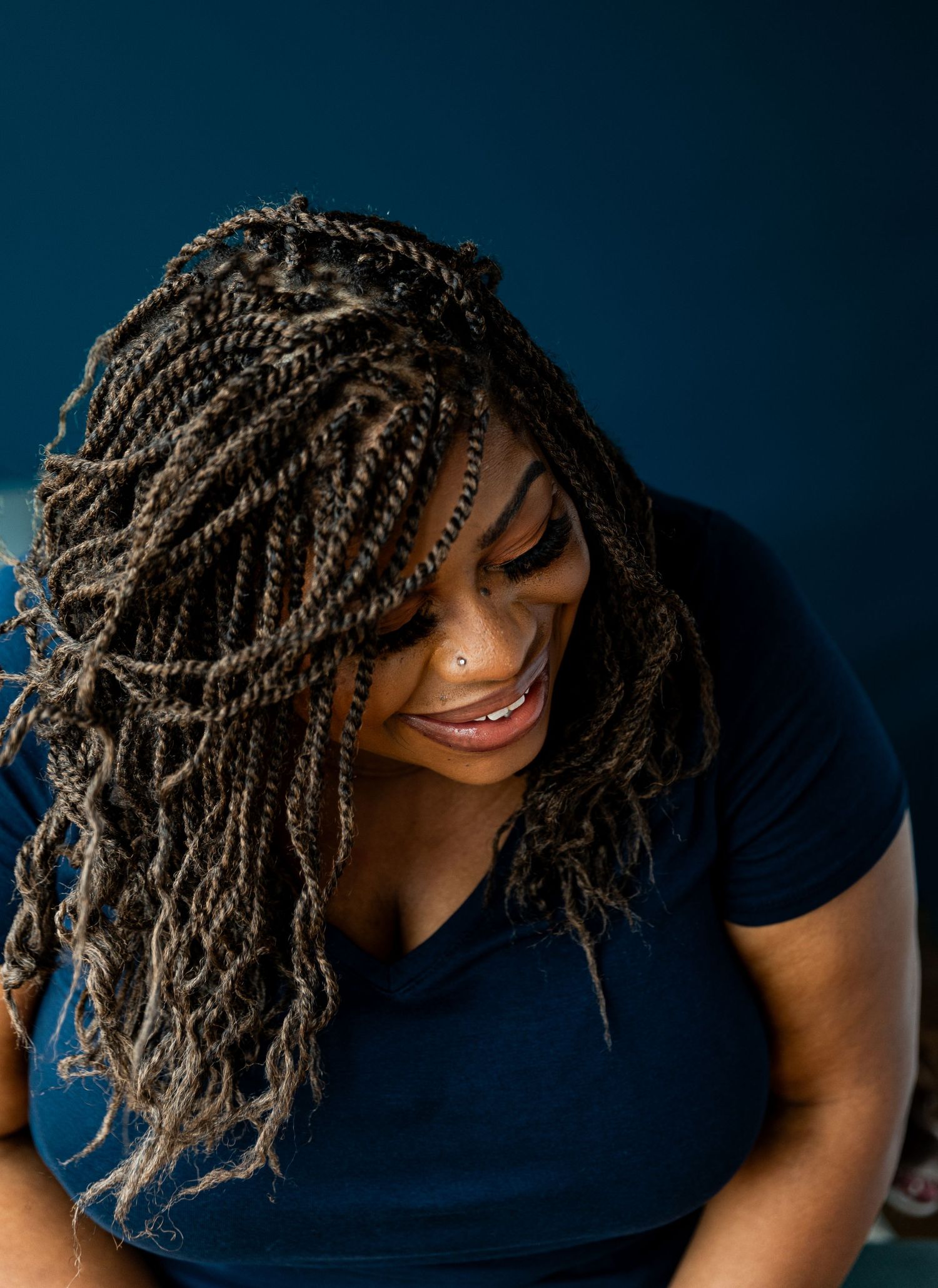 A Black woman with long twisted braids smiles downward, wearing a navy blue top against a dark teal background.
