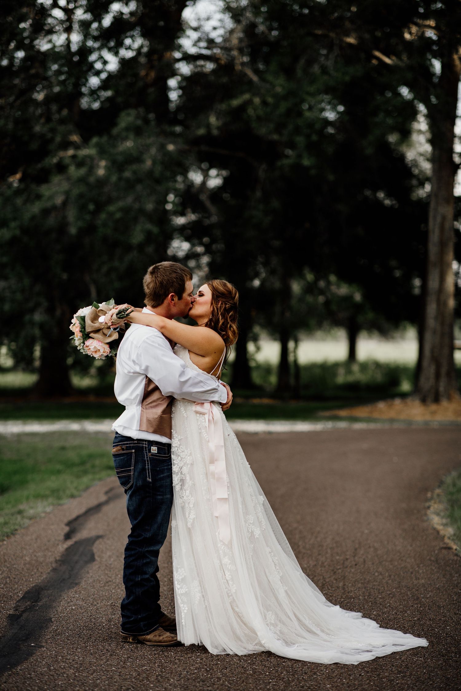 Bride and groom sharing a romantic kiss on a tree-lined road outdoors.