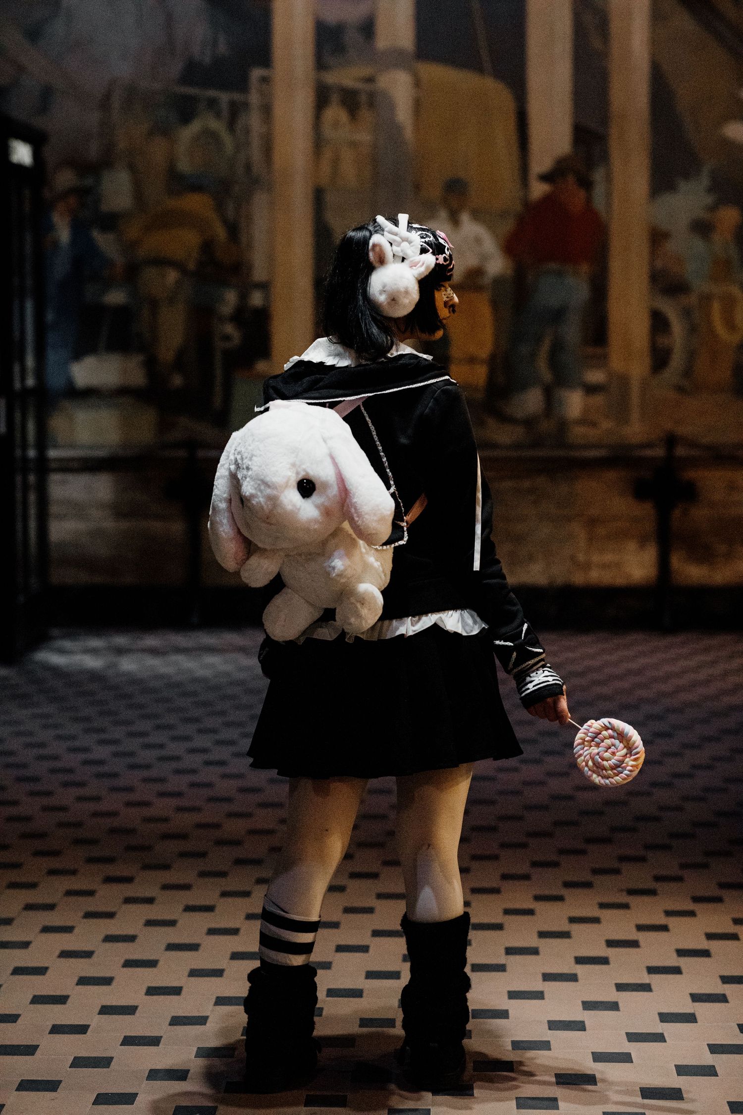 Gothic girl in black outfit holding lollipop with white bunny plush backpack, standing in ornate interior.