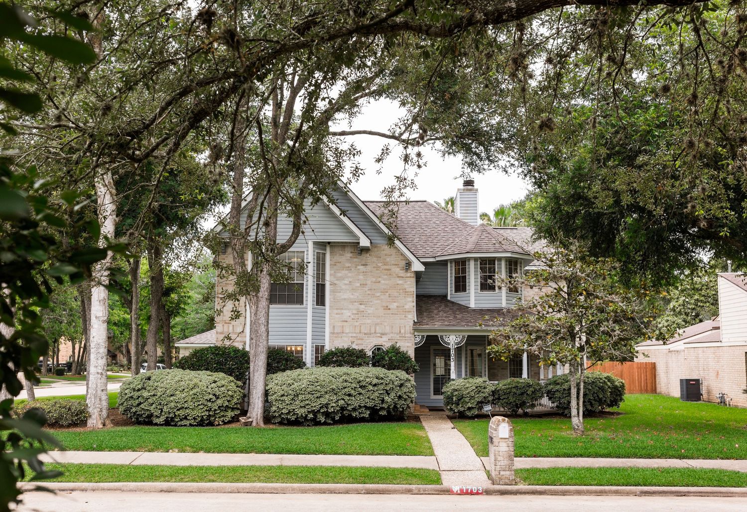 Two-story stone and siding home with wrap-around porch, lush landscaping, and mature trees lining a quiet suburban street.