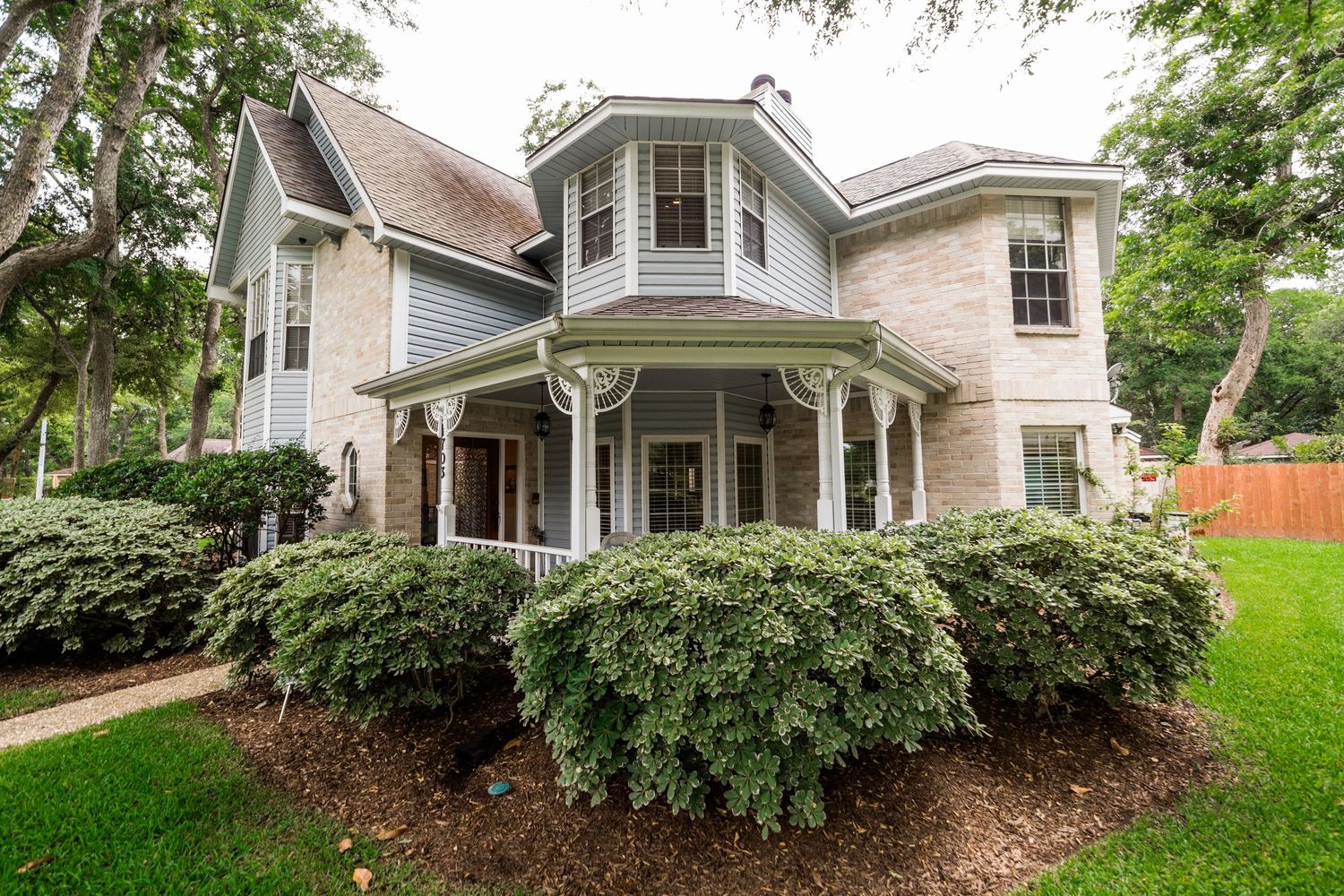 Two-story Victorian-style brick home with bay window, covered porch, and lush green landscaping surrounded by mature trees.