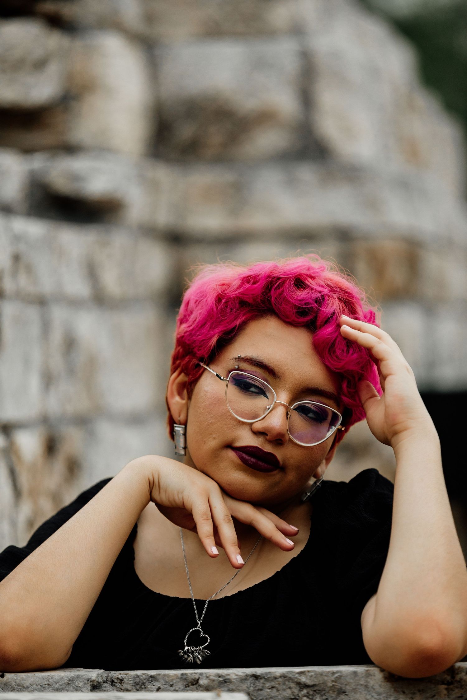 Young woman with pink curly hair and glasses rests her chin on hands, wearing black top and silver necklace outdoors.