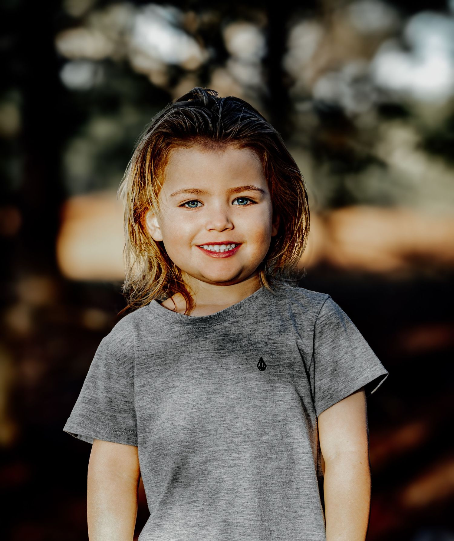 Smiling young child with blue eyes wearing gray Volcom tee in warm outdoor bokeh background.