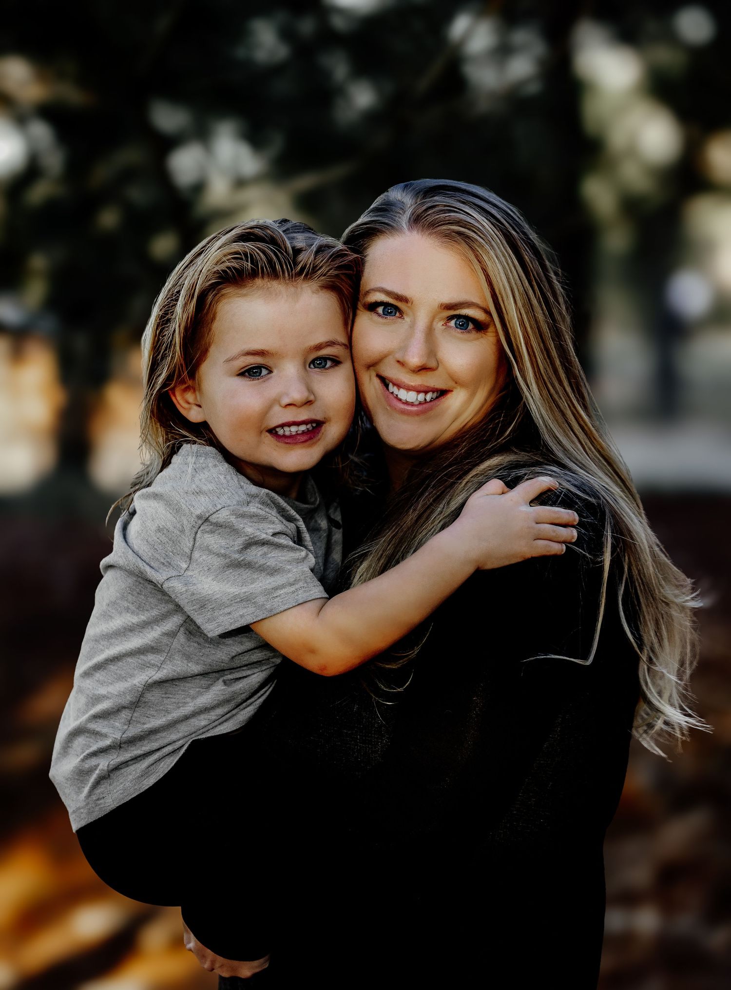 A smiling woman holds a happy young boy who wraps his arms around her in a warm outdoor portrait.