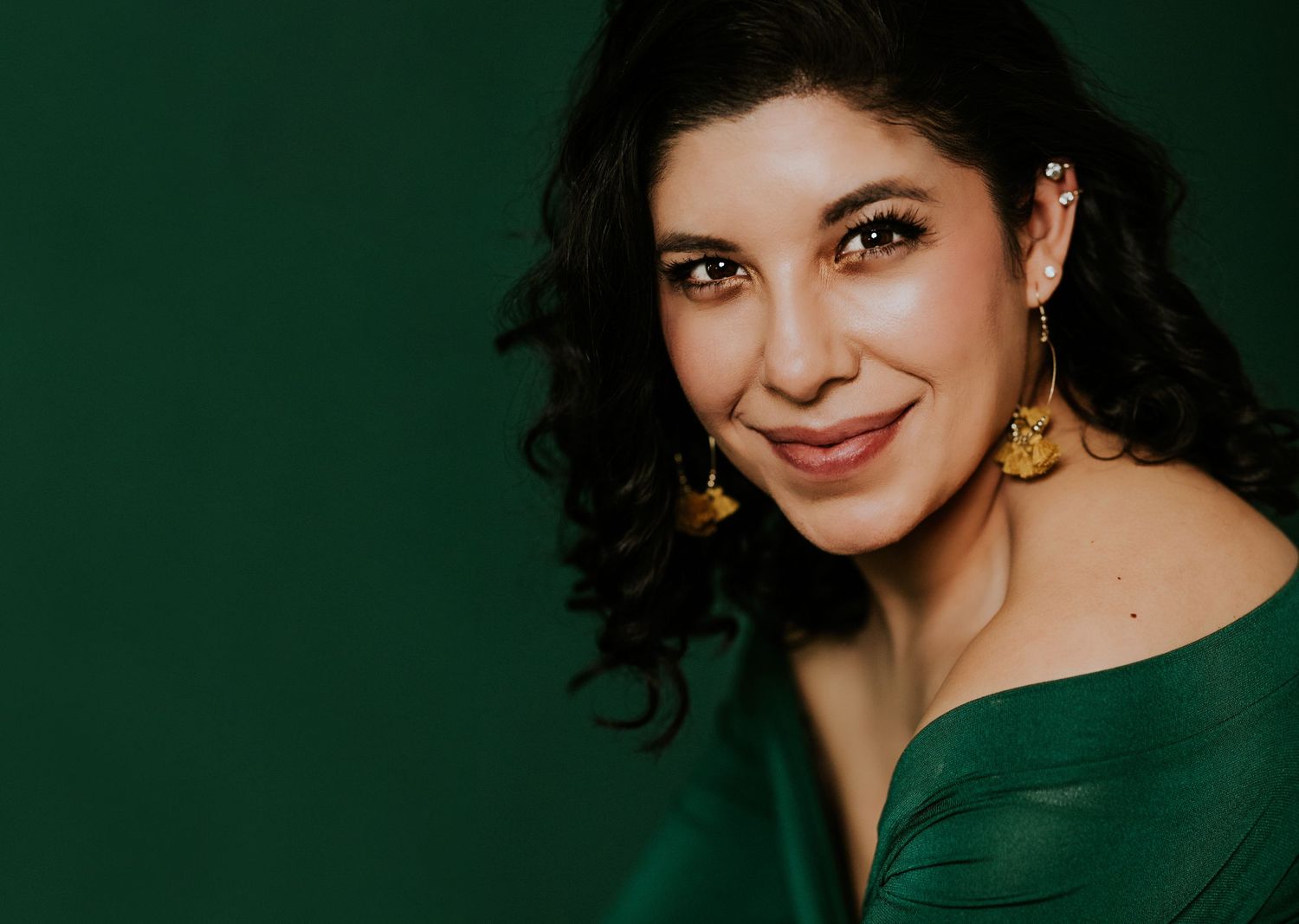 Woman in green dress with gold earrings smiling against dark green background.