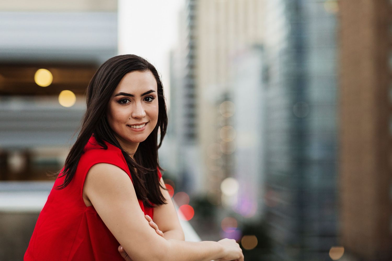 Young woman in red dress smiling on urban rooftop with city skyscrapers in blurred background.